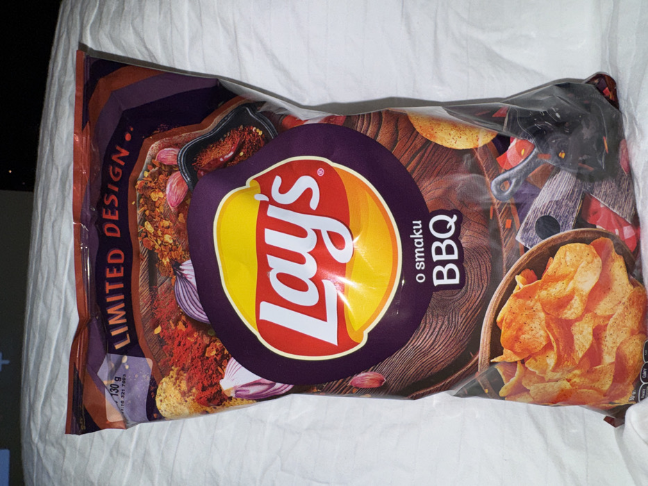 A crumpled bag of Lay's BBQ flavored potato chips rests on a white surface. The bag boasts a "Limited Design" with images of spices, onions, and a wooden bowl overflowing with chips. The Lay's logo is prominently displayed, along with the "o smaku BBQ" text. The background features a dark wood pattern with BBQ equipment like a spatula and cutting board. The bag appears partially open.