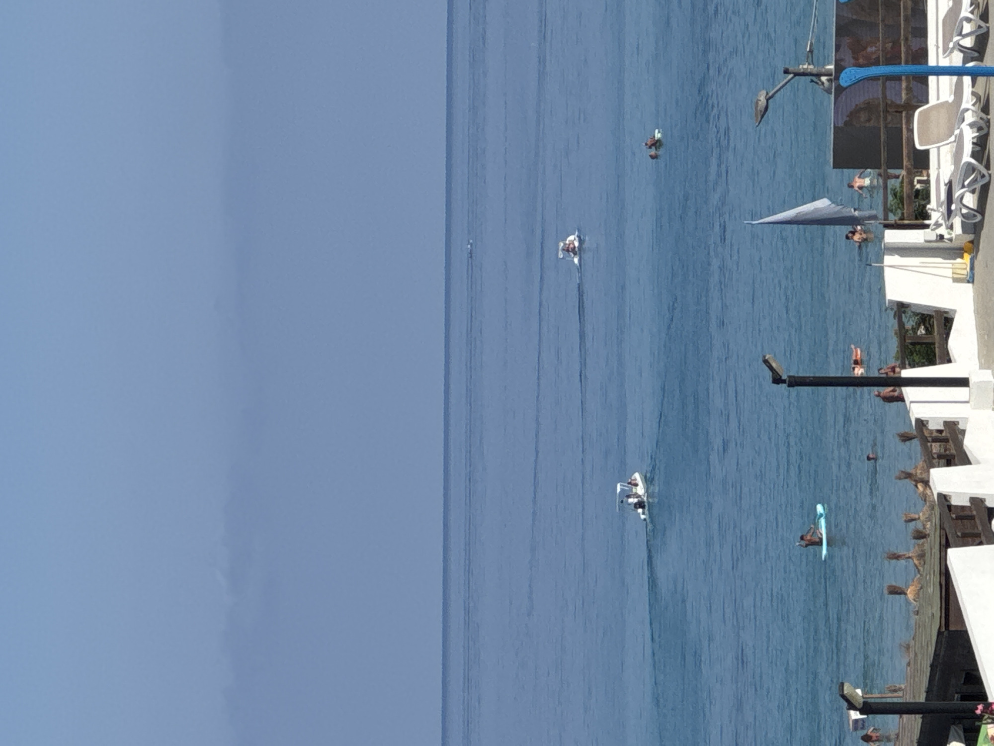 The image depicts a coastal scene with the Tyrrhenian Sea in Capaci, Italy. The water is a vibrant blue, with several small boats and people enjoying various activities. In the mid-ground, a white motorboat with several occupants is navigating the water. Further out, another, smaller boat is visible. Closer to the shore, a person is paddleboarding. On the far right of the image, lounge chairs and a flag suggest a resort or beach area. The sky is a pale blue, with some atmospheric haze obscuring the distant horizon.