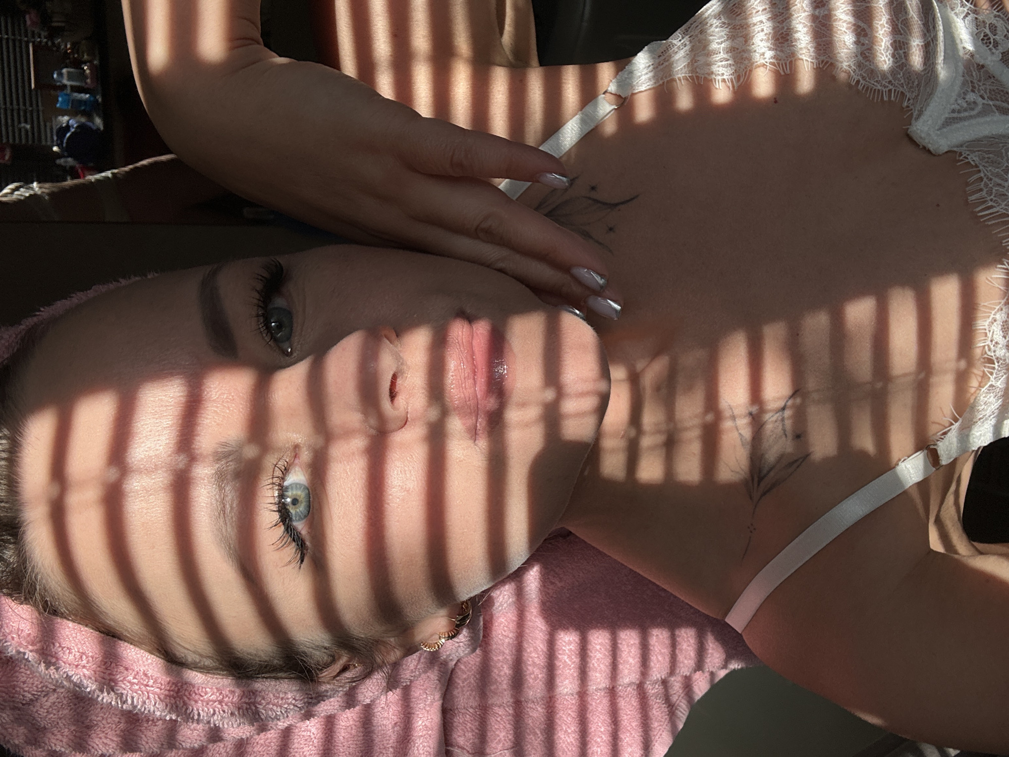 A woman is lying on her back, possibly in a bed covered with a pink towel. Her face is partially obscured by shadows of blinds. She is wearing a white top with lace trim and has a floral tattoo on her upper chest. Her hand rests on her chest, near her collarbone. The location appears to be indoors and possibly in a residential setting, but there are no landmarks or indicators connecting it specifically to the city of Chaikovsky, Russia.