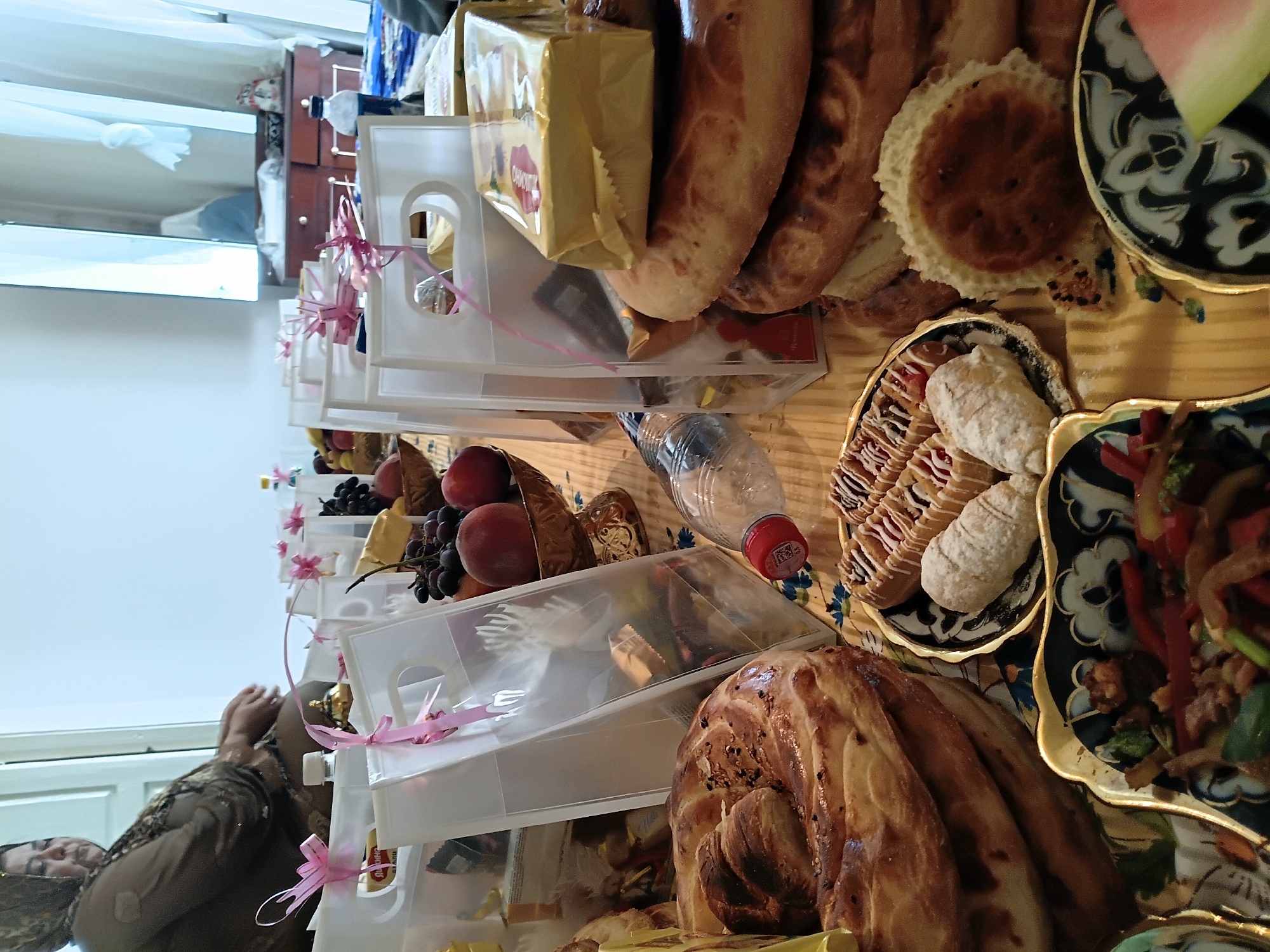 The image depicts a close-up, eye-level view of a table laden with food and gift bags, likely set up for a gathering or celebration. In the background, a woman wearing a headscarf and a traditional patterned dress is partially visible, seated and looking down. The table is covered with a yellow and white striped cloth.
Dominating the foreground are various baked goods, including large, round, braided breads with sesame seeds and smaller, flatter bread pieces. A decorative plate holds several pastries, some topped with icing and jam, and others appear to be crescent-shaped and dusted with powdered sugar. Another ornate plate in the lower left corner contains a colorful dish of stir-fried vegetables with meat. To the right, a slice of watermelon is visible on a dark patterned plate.
Scattered throughout the scene are several clear plastic gift bags, many tied with pink ribbon. These bags contain various items, including what appear to be chocolates, candies, and possibly other small gifts. A plastic water bottle with a red cap is positioned in the center of the frame. In the middle ground, a bowl filled with grapes and peaches is also visible. The overall impression is one of abundance and preparation for an event. The image appears to have been taken indoors, with natural light filtering in from a window or doorway on the left, illuminating the scene. The presence of a dresser with decorative items and curtains in the background suggests an indoor domestic setting, possibly in the city of Osh, Kyrgyzstan, given the cultural context hinted at by the woman's attire and the food items.
