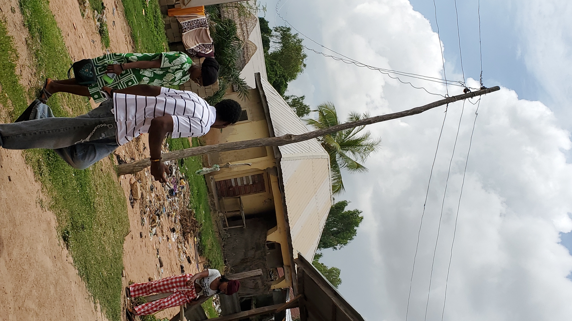 In Jalingo, Nigeria, under a cloudy sky, three individuals are visible in a dusty, rural setting. A person in a white and black striped shirt and blue jeans walks away from the camera, holding a mobile phone. Beside them, a woman in a green and white patterned dress and a black headscarf walks in the same direction, carrying a purse. To the right, a woman in a red and white checkered outfit stands, facing away from the camera. Behind them, a yellow building with a corrugated metal roof and wooden doors sits on a plot littered with debris. Clothes are hung on a line to the left, and a wooden utility pole with wires runs through the center of the frame. Palm trees and other greenery are scattered throughout the scene.