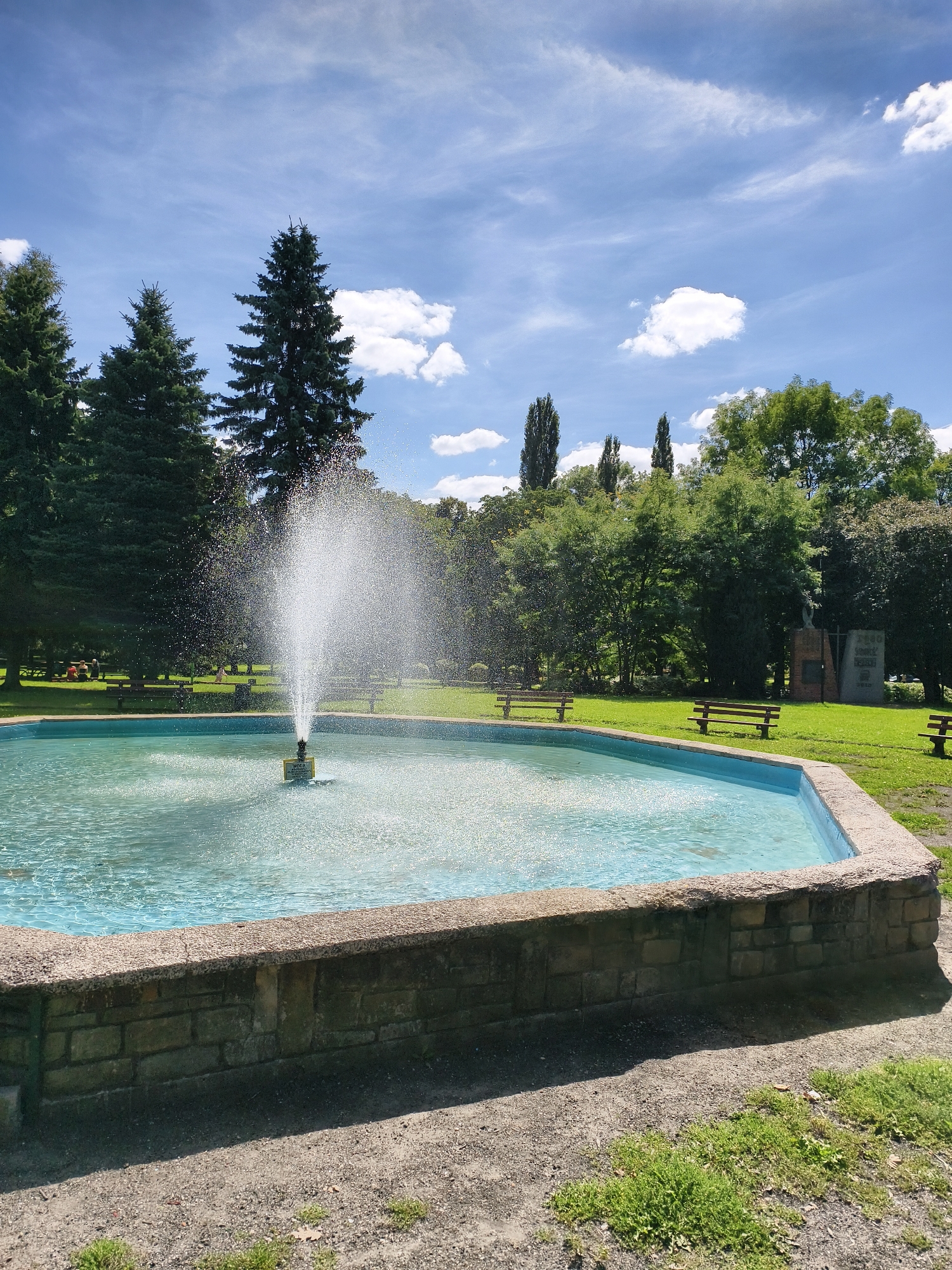 The image depicts a sunny day in a park with a fountain in the foreground. The fountain is active, spraying water upwards in a circular pattern into a light blue pool. The pool has a stone and brick border. Behind the fountain, a lush green lawn is visible, dotted with several wooden benches. In the background, a variety of trees, including tall conifers and deciduous trees, create a dense treeline. Further back, a statue or monument is partially visible, along with a stone structure. The sky is blue with scattered white clouds. The overall impression is a peaceful, recreational outdoor space. This location is the Park Miejski in Tczew, Poland.