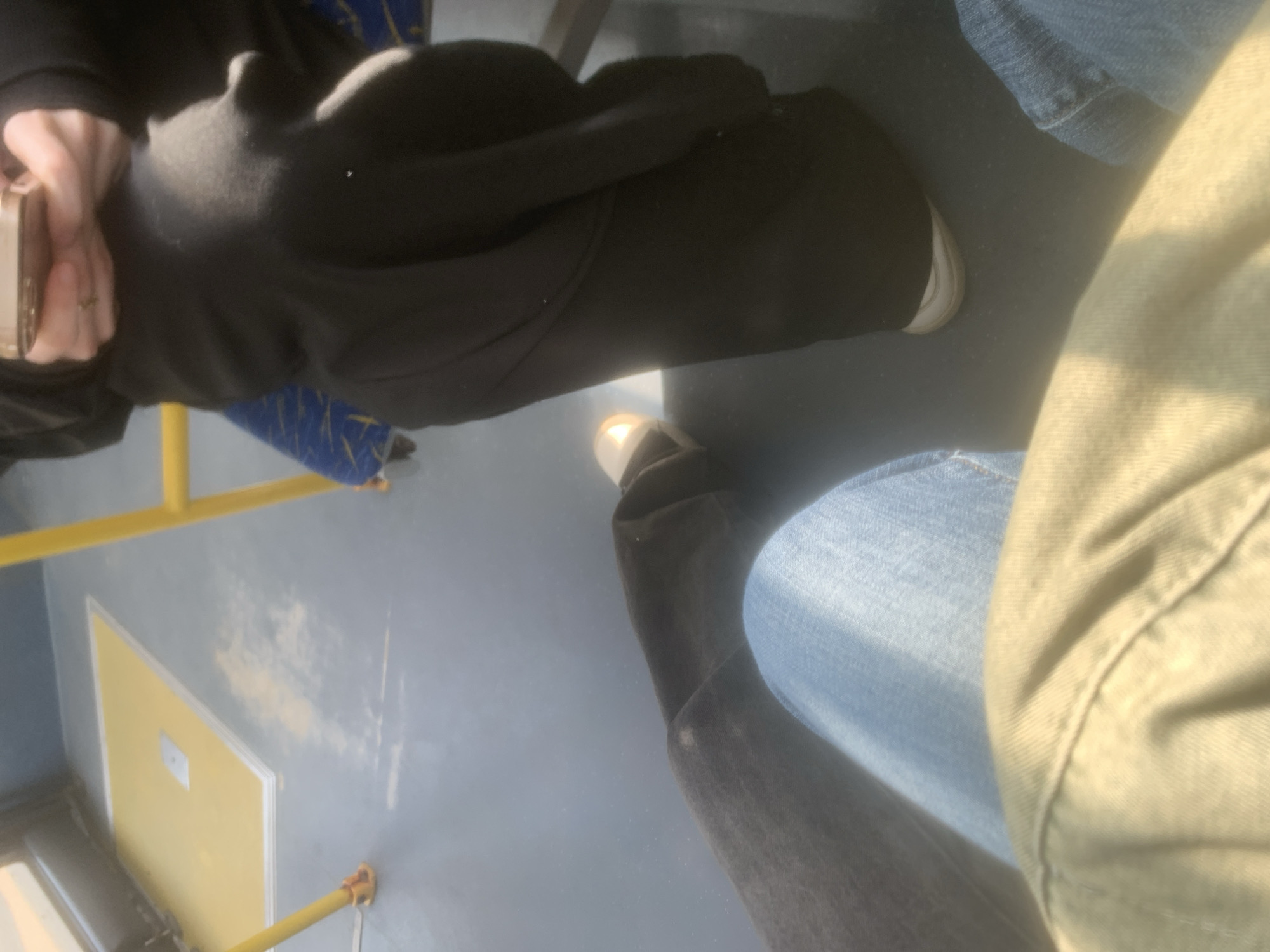 The image depicts the interior of a public transport vehicle, likely a bus or tram, seen from a low angle. Two individuals are partially visible, primarily their legs and lower torsos.
On the left side of the frame, a person wearing dark trousers or sweatpants and white sneakers is holding a light-colored, possibly golden or rose-gold, rectangular object, which appears to be a phone or wallet. A ring is visible on one of their fingers. Behind this person, a blue seat with a yellow geometric pattern can be seen.
On the right, closer to the camera, is another person wearing blue jeans and light-colored shoes. Part of a light green or beige fabric, possibly their clothing, is also visible.
The floor of the vehicle is light grey. Yellow grab bars or poles, characteristic of public transport, extend across the bottom and left side of the image. On the vehicle's wall, a rectangular yellow panel with a white border is present, and above it, some faded white or yellowish marks are scattered on the grey wall surface.
Bright sunlight streams into the vehicle, creating strong highlights and shadows, suggesting it is daytime with clear weather outside. No specific activity other than being transported is apparent. No legible text is visible within the frame.