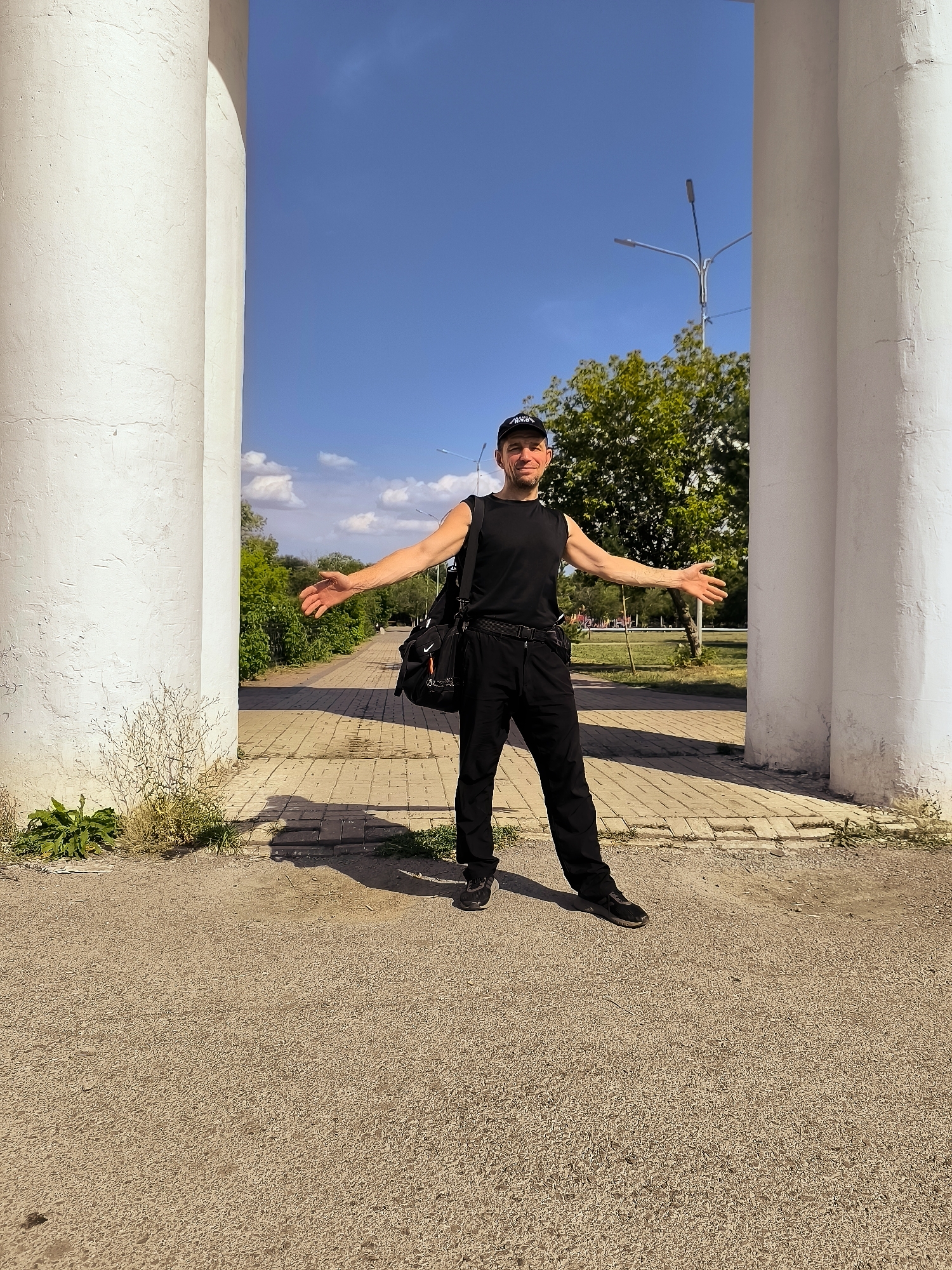 A man in a black sleeveless shirt, black pants, a black baseball cap, and dark shoes stands outdoors under a bright blue sky with scattered clouds. He is positioned between two large, fluted white columns, with his arms outstretched to his sides. A black messenger bag with orange and grey accents hangs across his left shoulder. He appears to be smiling and looking towards the camera.
The ground in front of the columns is a light-colored gravel surface. Behind the columns, a paved walkway with a brick pattern extends into the distance, flanked by green foliage and trees. A street lamp with multiple lights is visible in the background, along with what appears to be a park or public space. The overall setting suggests an outdoor park or entrance area in a city.
The image does not contain any specific visual cues that would definitively place the location in the city administration of Qarağandı, Kazakhstan. The environment is generic enough to be found in many urban or park settings.