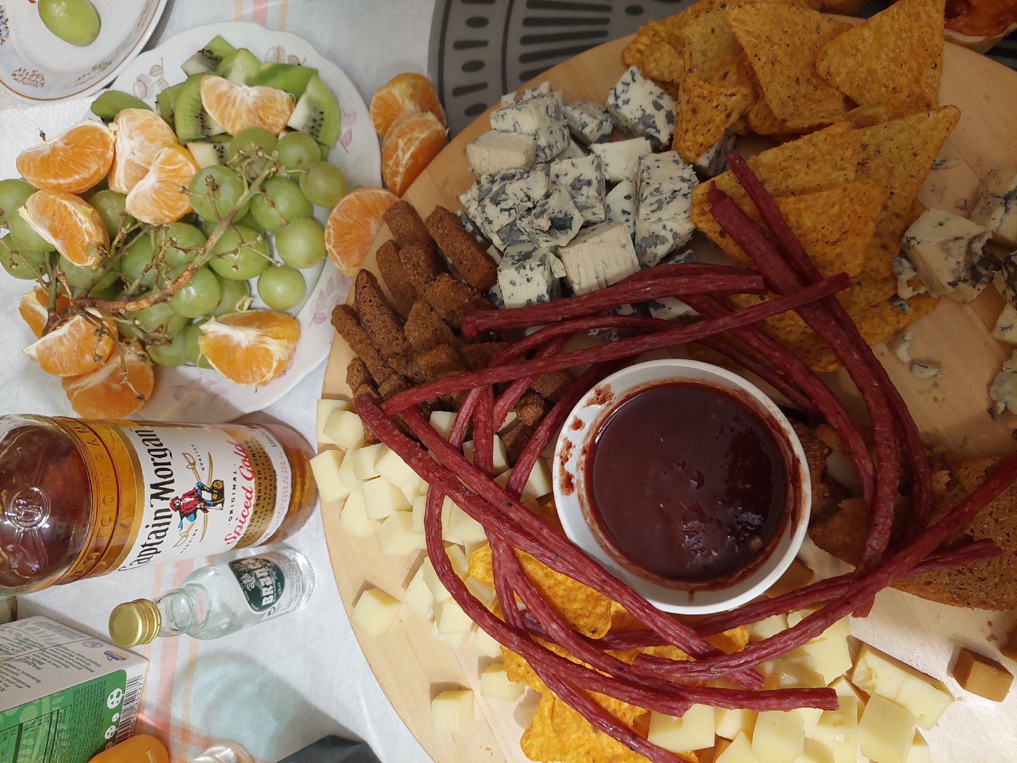 The image shows a charcuterie board and a fruit plate. The charcuterie board includes blue cheese, cheddar cheese cubes, bread sticks, sausage sticks, nachos, and a bowl of dark sauce, possibly a jam. The fruit plate has grapes, tangerine slices, and kiwi slices. There is also a bottle of Captain Morgan Spiced Gold rum and a small bottle of BranYa vodka.
The location is not mentioned in the image. It cannot be confirmed from the image alone if it is in Ternopil, Ukraine or any other city.