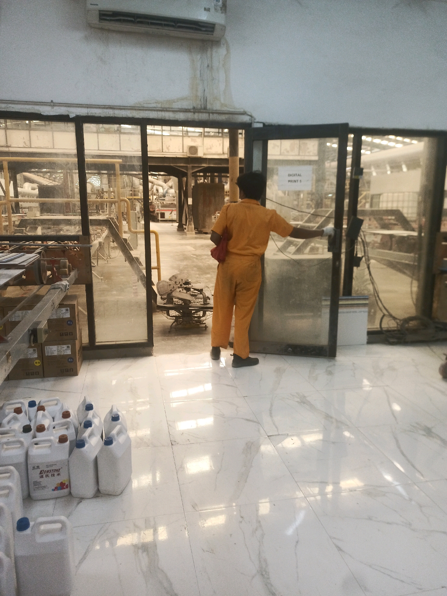 A person in orange coveralls is standing near a glass door in what appears to be an industrial or factory setting. The person is facing away from the camera and has their right hand extended, touching the door frame. They are also carrying a red bag over their left shoulder. The floor is tiled with a marble pattern. In the foreground, there are several white plastic containers with blue and brown caps. In the background, through the glass, there is industrial equipment and machinery. An air conditioning unit is visible on the wall above the doorway. The scene is lit by overhead lights, casting reflections on the floor. The overall impression is one of an active workplace.