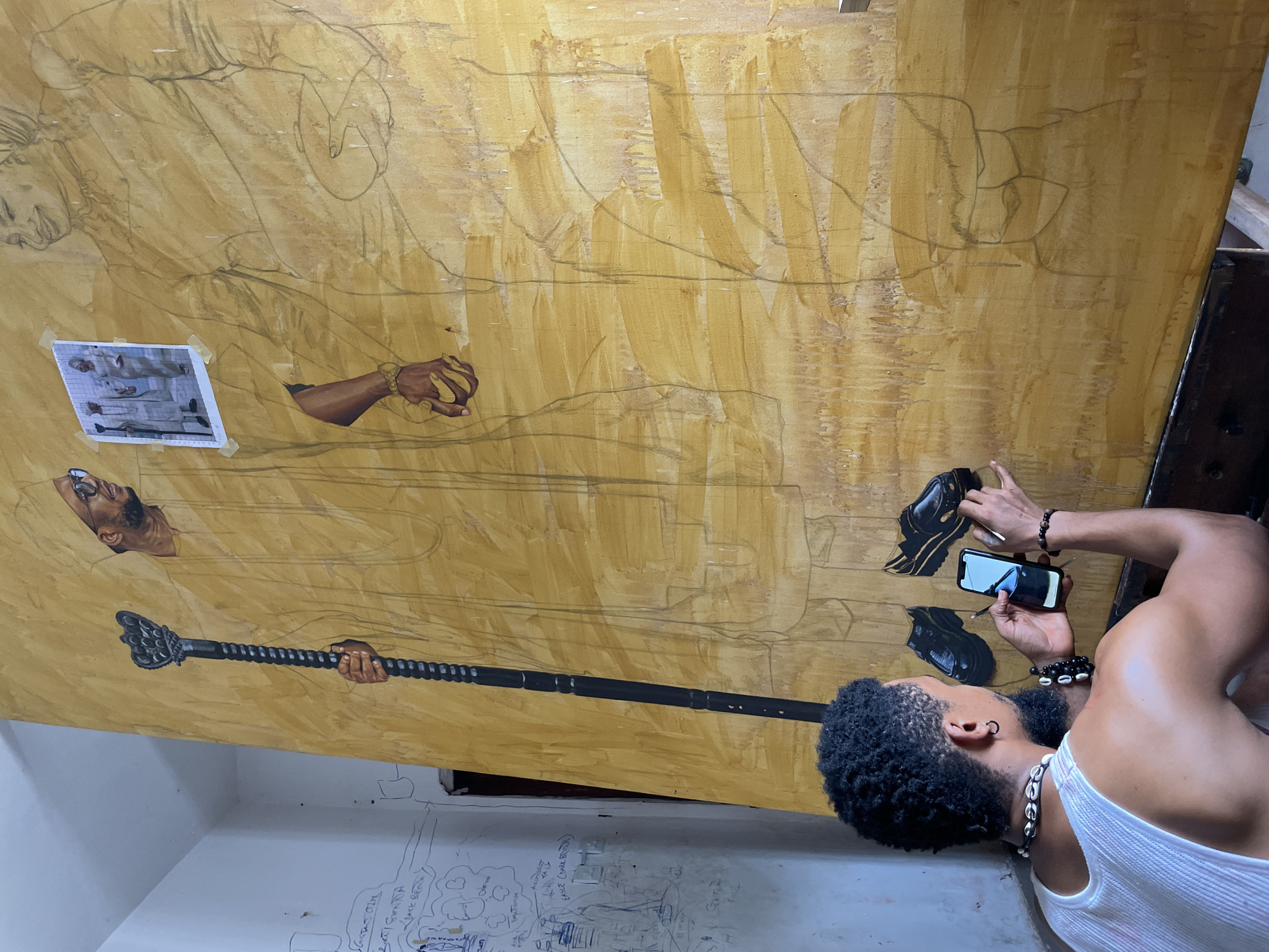 An artist is captured in the process of creating a large-scale painting, which is propped upright in what appears to be a studio setting. The main subject is a man, seen from behind and the side, with dark, curly hair and a beard. He wears a white tank top, hoop earrings, a necklace made of cowrie shells, and several beaded and cowrie-shell bracelets on his right wrist. He is holding a smartphone in his left hand, its screen reflecting light, and his right hand is positioned near the artwork, suggesting he is actively painting or adjusting something.
The artwork itself is a large canvas or board, featuring an underpainting of warm yellow and light brown tones with visible brushstrokes. Over this base, several figures are outlined in darker lines, indicating an initial sketch phase for a multi-figure composition. Parts of the painting are already detailed: a dark-skinned hand gripping a black, ornate staff or mace is prominent in the lower left, along with a dark-skinned head wearing sunglasses further up. Another dark-skinned arm with a gold bracelet is visible higher on the canvas. Two black, formal-looking shoes are painted near the artist's hands, appearing almost complete. A small printed reference photograph, showing two figures in white garments, is taped onto the canvas.
Below the artwork, on a white wall, faint pencil sketches and illegible text are visible, characteristic of an artist's workspace. The overall scene conveys a sense of focused artistic creation. The given context indicates this scene takes place in Ado, Nigeria.