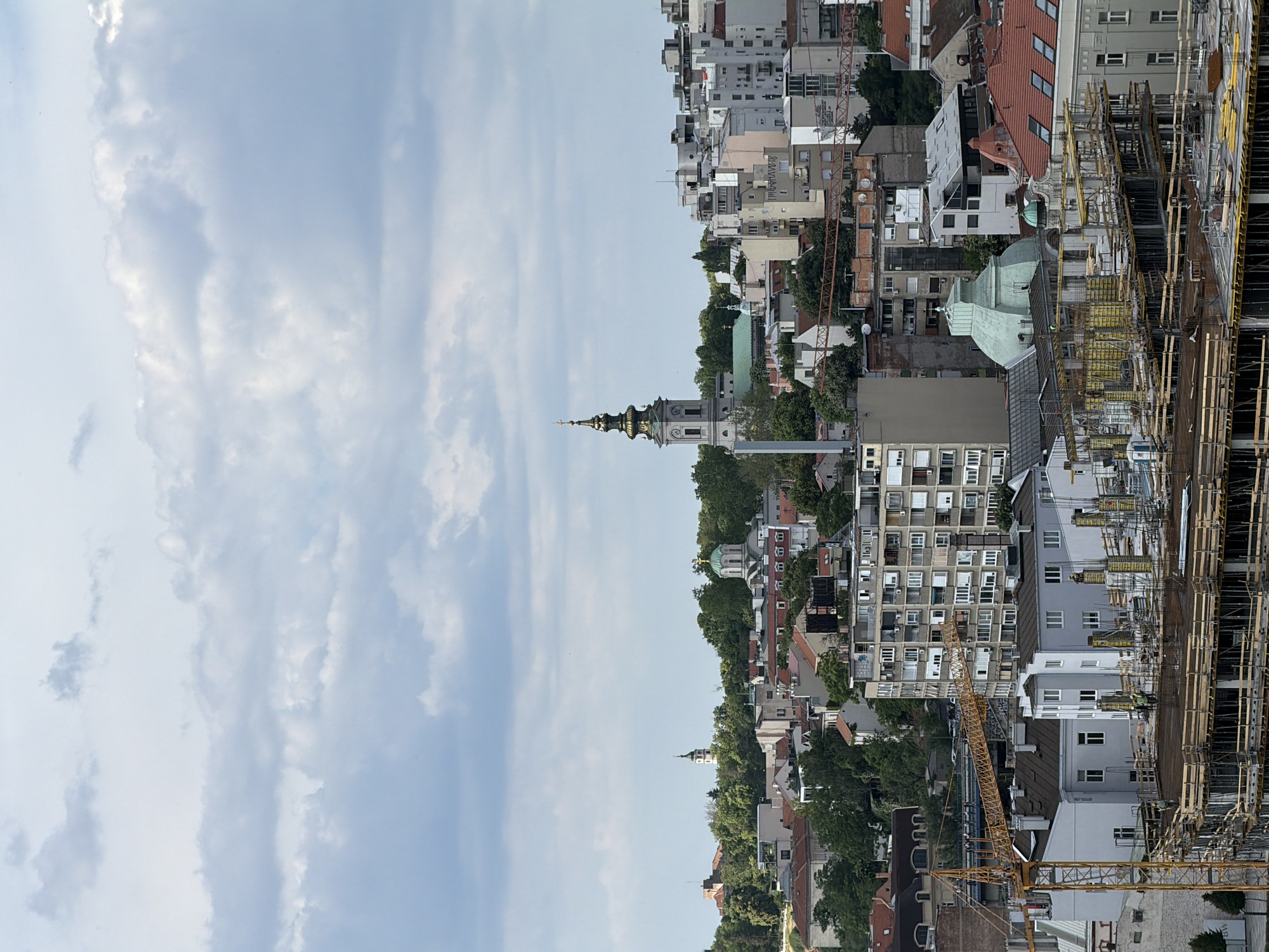 A wide-angle shot of Belgrade, Serbia, from an elevated perspective shows a skyline dominated by buildings, a church spire, and a construction site. The sky is light blue with scattered clouds. The architecture consists of mixed-height buildings, ranging from residential structures to high-rises, with various roof types, including sloped and flat. The church spire is prominent, painted light and topped with a gold dome. A construction site is visible on the right side of the image, with yellow cranes and scaffolding. Trees are dispersed throughout the cityscape.