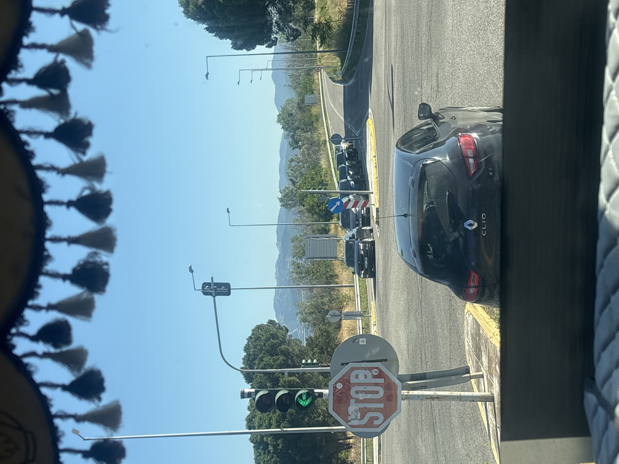 The image captures a view from inside a vehicle, looking out at a road intersection in Kikidio, Greece. In the foreground, a dark-colored hatchback, identified by the "CLIO" badge on its rear, is parked, partially obscuring the view. Above the road, a decorative fringe with tassels hangs from the vehicle's interior, casting shadows.
The intersection features a prominent stop sign, octagonal and red with white lettering, positioned on a metal pole. Adjacent to the stop sign is a traffic light displaying a green left-turn arrow. Another traffic light is visible further down the road, displaying a green light. Several cars are lined up at the intersection, waiting at the traffic light. The road curves to the right, leading into the distance.
The environment is characterized by a clear blue sky and a bright, sunny day. Trees and vegetation line the sides of the road, and in the background, mountains are visible under the expansive sky. Streetlights are present at various points along the road, indicating an urban or suburban setting. The overall scene suggests a typical day at a Greek road intersection.