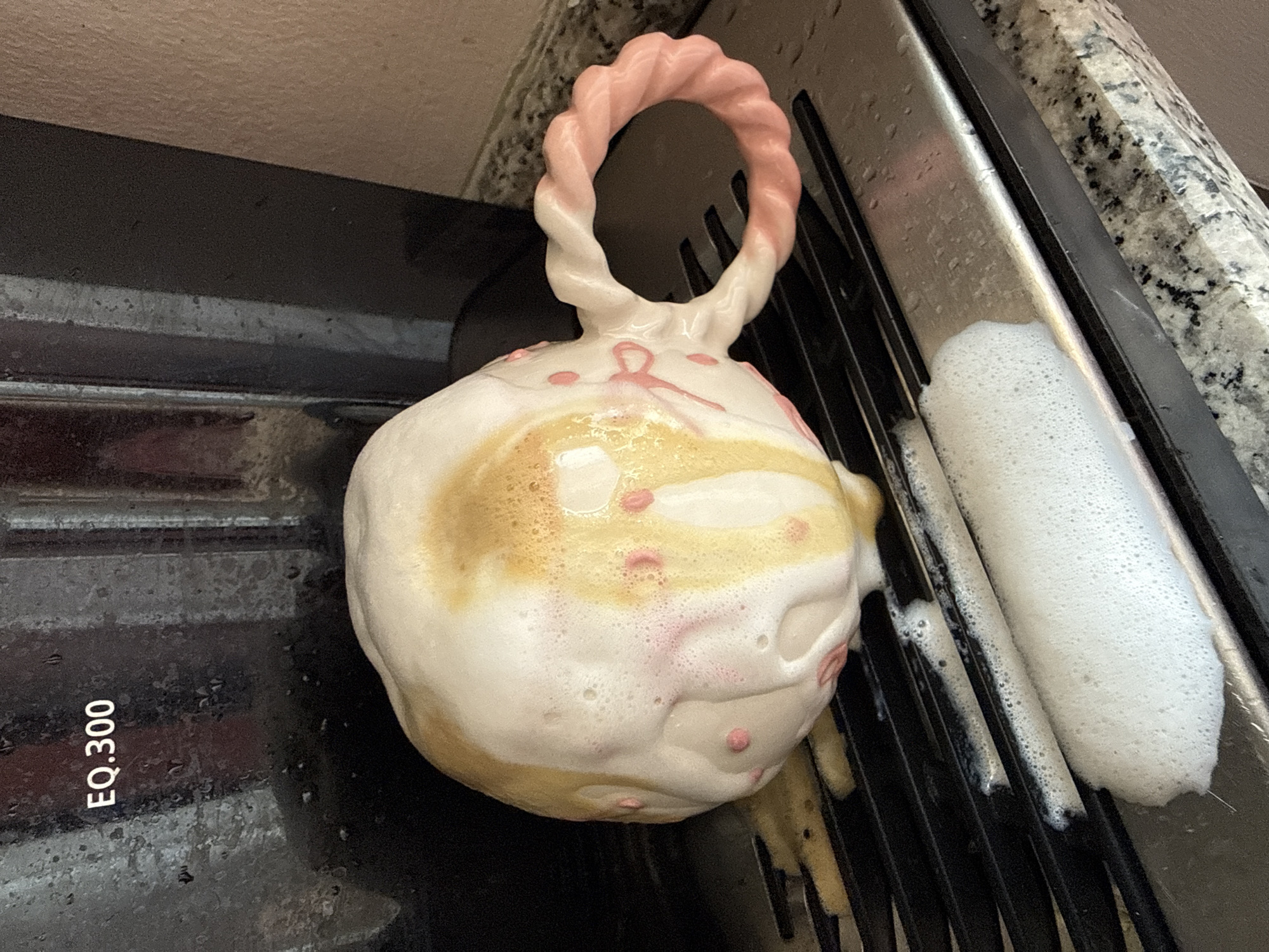The image shows a close-up of a white and pink ceramic mug that has tipped over and is covered in foam, likely from a coffee or milk-based drink. The mug has small pink dots and pink ribbon-like decorations. It is resting on a black metal grate, which appears to be part of a coffee machine's drip tray. There is a significant amount of foam and liquid on the grate and overflowing onto the counter. In the background, a portion of a dark-colored coffee machine is visible, with the text "EQ.300" displayed. The surrounding counter has a speckled granite pattern. The scene is indoors, likely in a kitchen or cafe setting. There is no indication of the city of Konya, Turkey, in the image.