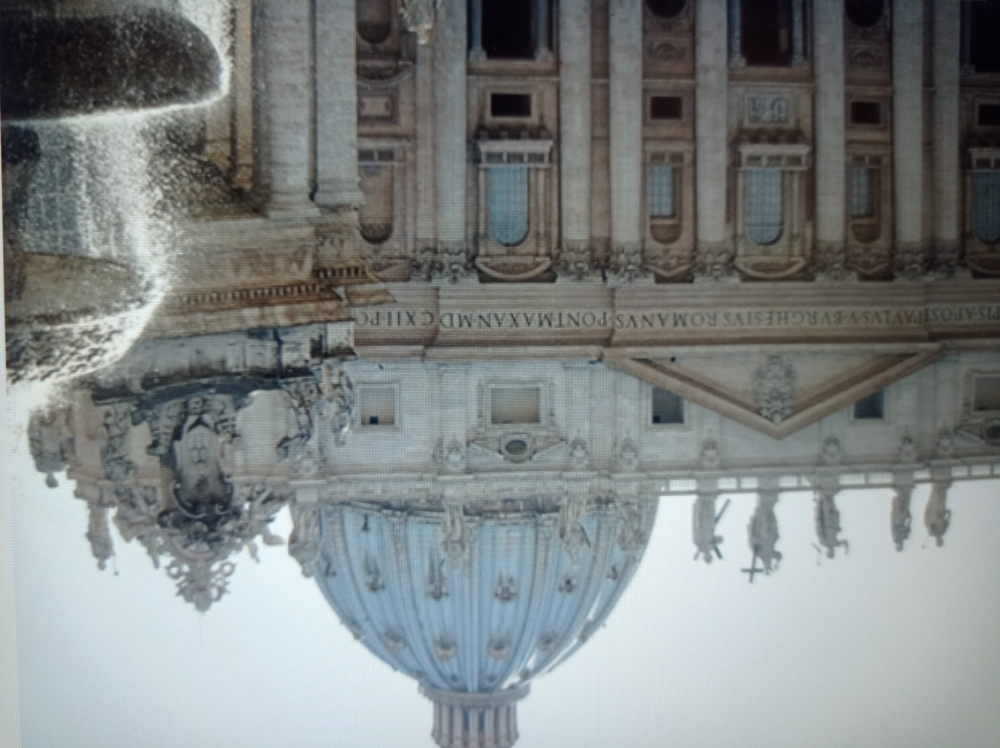 The image shows a grand architectural structure, likely a basilica or cathedral, with a prominent dome and ornate facade. The building features classical columns, arched windows, and numerous statues adorning its exterior. In the foreground, to the right, a fountain is visible with water cascading down its tiered structure. The sky is overcast, suggesting a diffused lighting condition. The inscription on the building reads "PIVS.APOST.PAVLVS.EVRGHESIVS.ROMANVS.PONT.MAX.AN.MDCXII." The presence of this inscription, which references Pope Paul V, indicates that the structure is St. Peter's Basilica in Vatican City, Rome, Italy, and not in Mukachevo, Ukraine. No people are clearly identifiable as subjects in the main composition, though there appear to be some statues and possibly distant figures. The scene is an outdoor architectural view focused on the grandeur of the building and the fountain.
