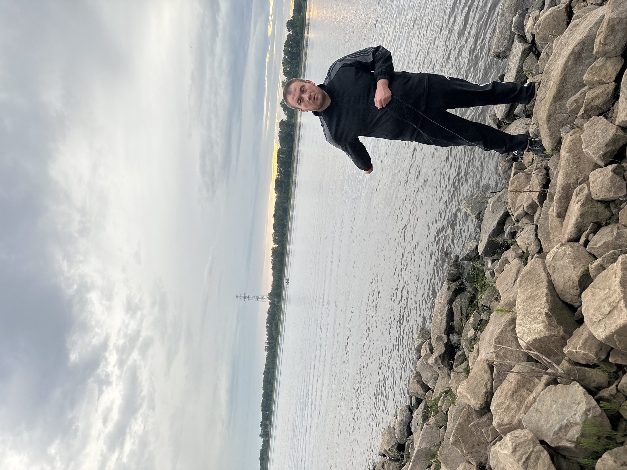 This is a landscape orientation, full-body, waist-up shot of a man standing on a rocky shoreline next to a body of water. The man is wearing a black Adidas tracksuit with gray stripes on the sleeves and pants. He is looking directly at the camera with a neutral expression. His right hand is clenched into a fist and held in front of him, while his left arm is bent at the elbow, with his hand resting near his chest.
The setting appears to be a river or a large lake, with a tree-lined shore visible in the distance. A large electrical pylon stands prominently on the opposite bank, suggesting a somewhat industrialized or developed area. The sky is overcast with dramatic clouds, and there is a hint of sunlight breaking through on the horizon, indicating either early morning or late evening. The water is calm, with ripples suggesting a gentle breeze. The rocks in the foreground are large and varied in size and shape, with some small green plants growing between them.
The overall mood is somewhat stark and solitary, with the man being the central focus against the natural landscape. The image is shot at an angle, making it appear tilted. There is no visible text in the image. The context provided indicates the location is Chaikovsky, Russia.