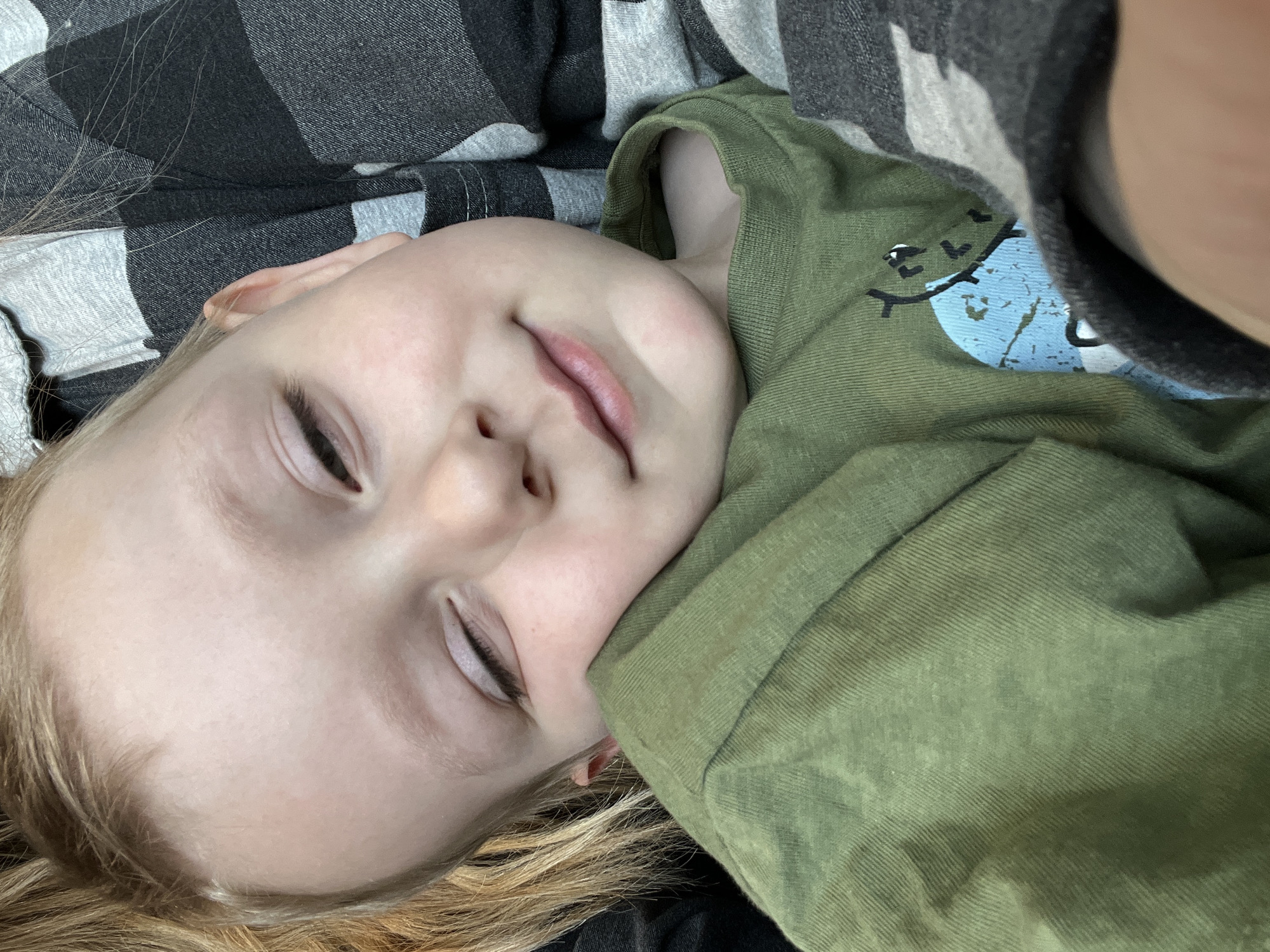 A close-up, eye-level portrait shows a young child, appearing to be asleep or resting, with their head gently tilted and resting against an unseen individual. The child has fair skin, light blonde hair, and closed eyes with visible eyelashes. A faint, soft smile is present on their lips. They are wearing a long-sleeved, olive-green t-shirt with a distinct vertical ribbed texture. A partial graphic design in light blue, black, and white is visible on the front of the shirt, depicting what appears to be part of a cartoon figure or animal. The unseen individual's shoulder and arm are visible, clad in a black and grey buffalo check patterned fabric, likely part of a shirt or blanket. The lighting is soft and even, suggesting an indoor environment. There are no visual cues within the image to indicate a specific geographic location, including the city of Moscow, Russia.