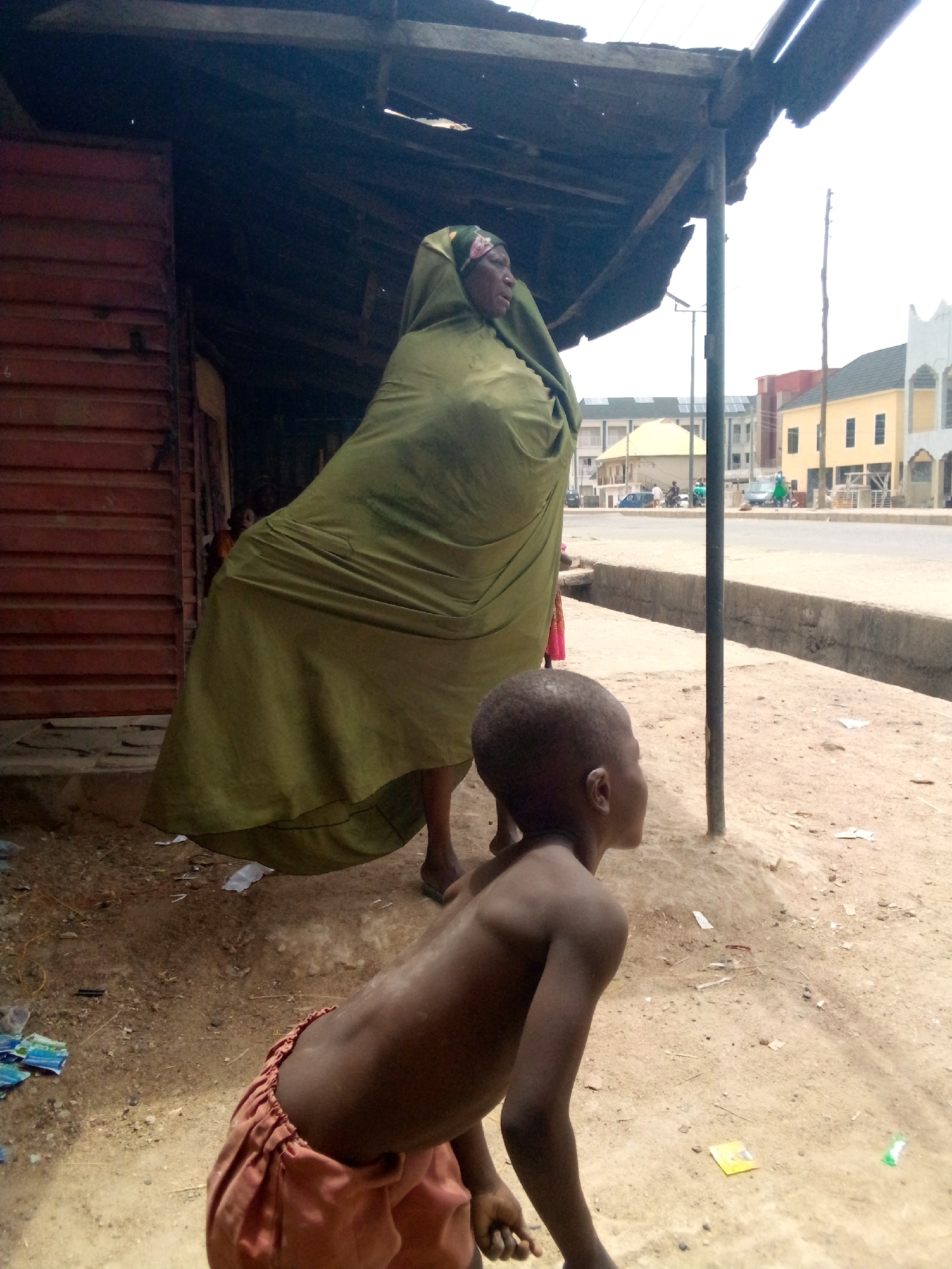 An eye-level, medium-shot captures an older woman draped in a large, green cloth and a young boy in front of a shop in the city of Minna, Nigeria. The woman, wearing a headscarf, stands under a weathered, wooden-roofed structure. The boy, with bare torso and wearing orange shorts, looks off to the side. The ground is dirt with scattered debris. The backdrop includes a street with buildings in the distance.