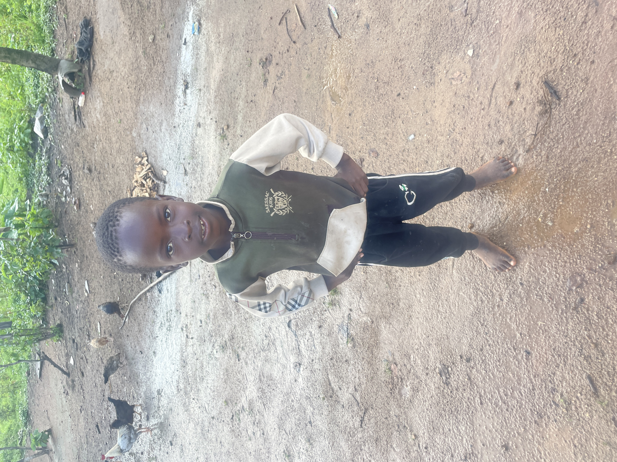 A young boy stands barefoot in a muddy, outdoor area in Jos, Nigeria. He is wearing a green and beige long-sleeved shirt with a zipper and a white pocket, and black pants with white stripes. The background shows dirt, scattered leaves, and several chickens. Lush green vegetation is visible in the upper part of the image.