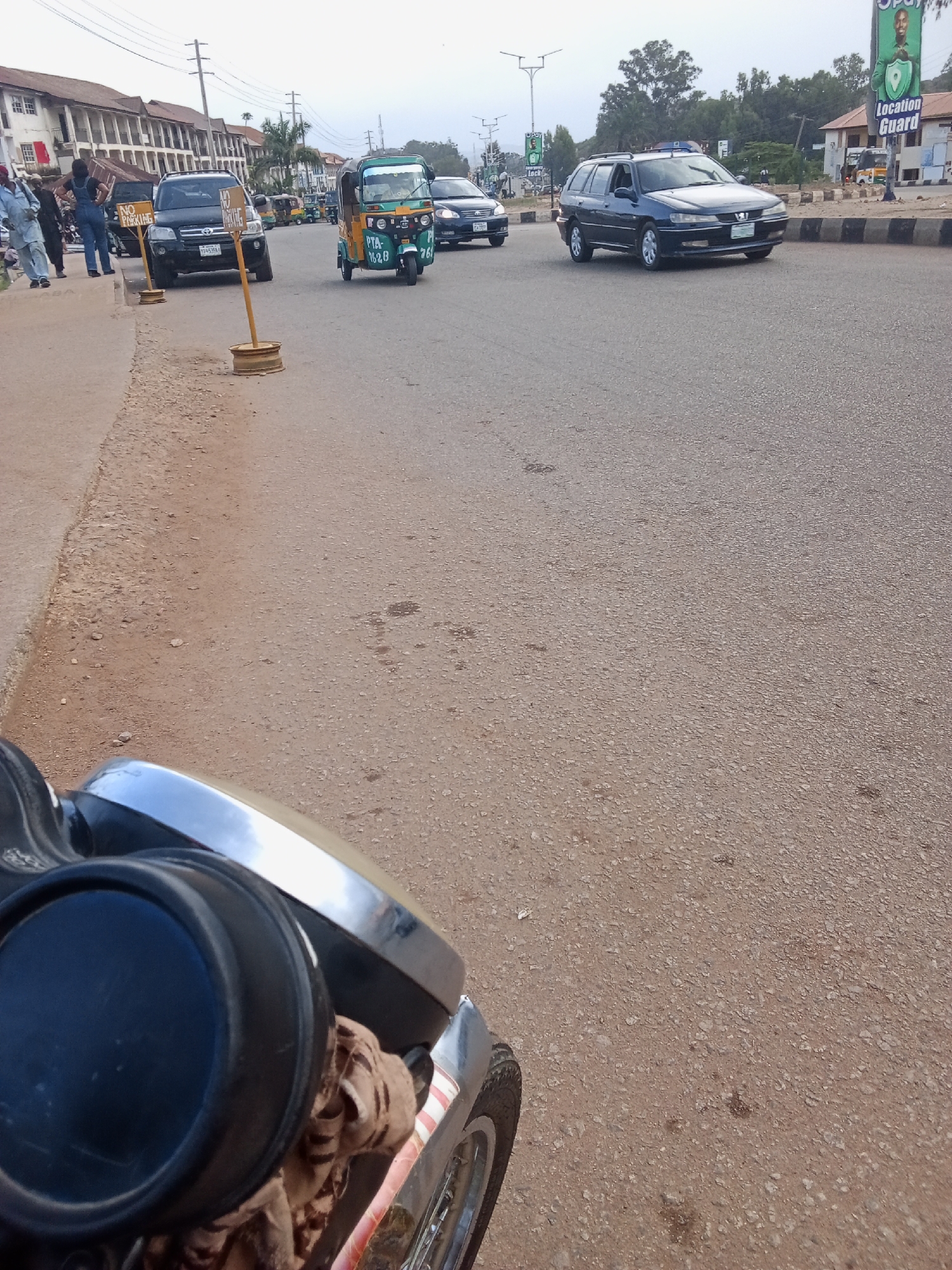 The image depicts a street scene in Gura Top, Nigeria. A motorbike is in the foreground, facing forward, with its headlight and part of the handlebars visible. The street is paved with asphalt and appears to be somewhat worn. In the background, several vehicles are present, including a blue station wagon, a black SUV, and a green and yellow auto-rickshaw. Pedestrians are visible on the sidewalk to the left of the road, where two "NO PARKING" signs are posted. Buildings line the left side of the street, and trees are visible in the distance. A billboard for "Location Guard" is also visible on the right side of the image. The sky is overcast.