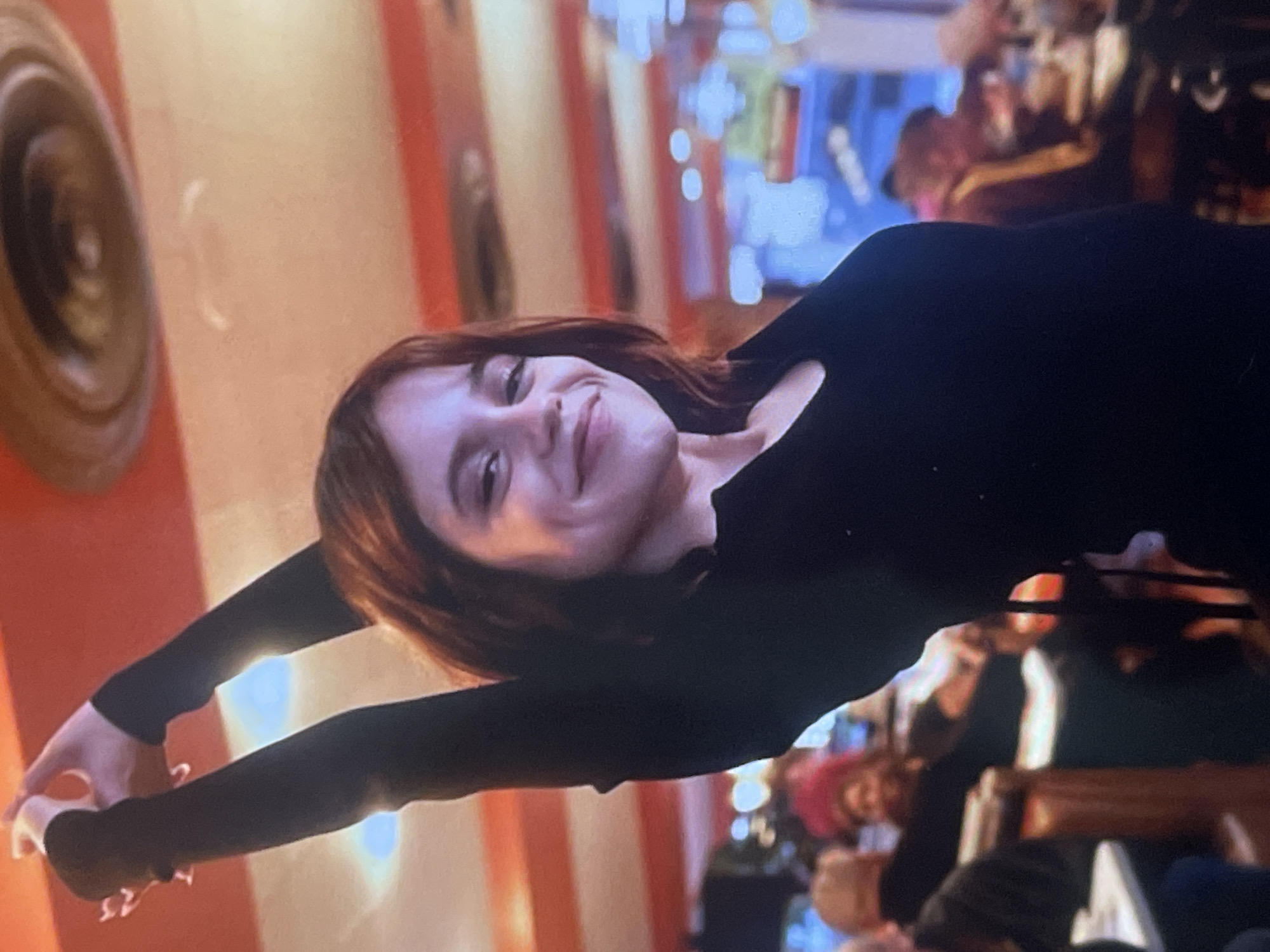 The image, rotated 90 degrees clockwise, prominently features a young woman smiling directly at the camera. She has reddish-brown hair and is wearing a black long-sleeved top. Her arms are extended outwards to her sides, suggesting a stretching motion or pose. Her expression conveys happiness or a playful mood.
The setting is indoors, appearing to be a restaurant or cafe, characterized by warm, artificial lighting. In the background, to the viewer's left (which would be the top when rotated correctly), a circular wall decoration, possibly a lamp or piece of art, is visible. The wall behind the woman is adorned with a distinctive pattern of alternating cream or white and orange vertical stripes. Further in the background, blurred figures of other people are discernible, along with indistinct shapes that suggest tables, chairs, and other light sources, indicating a bustling and social environment. The overall ambiance is lively and casual. There is no visible text in the image. This scene is located in Poltava, Ukraine.