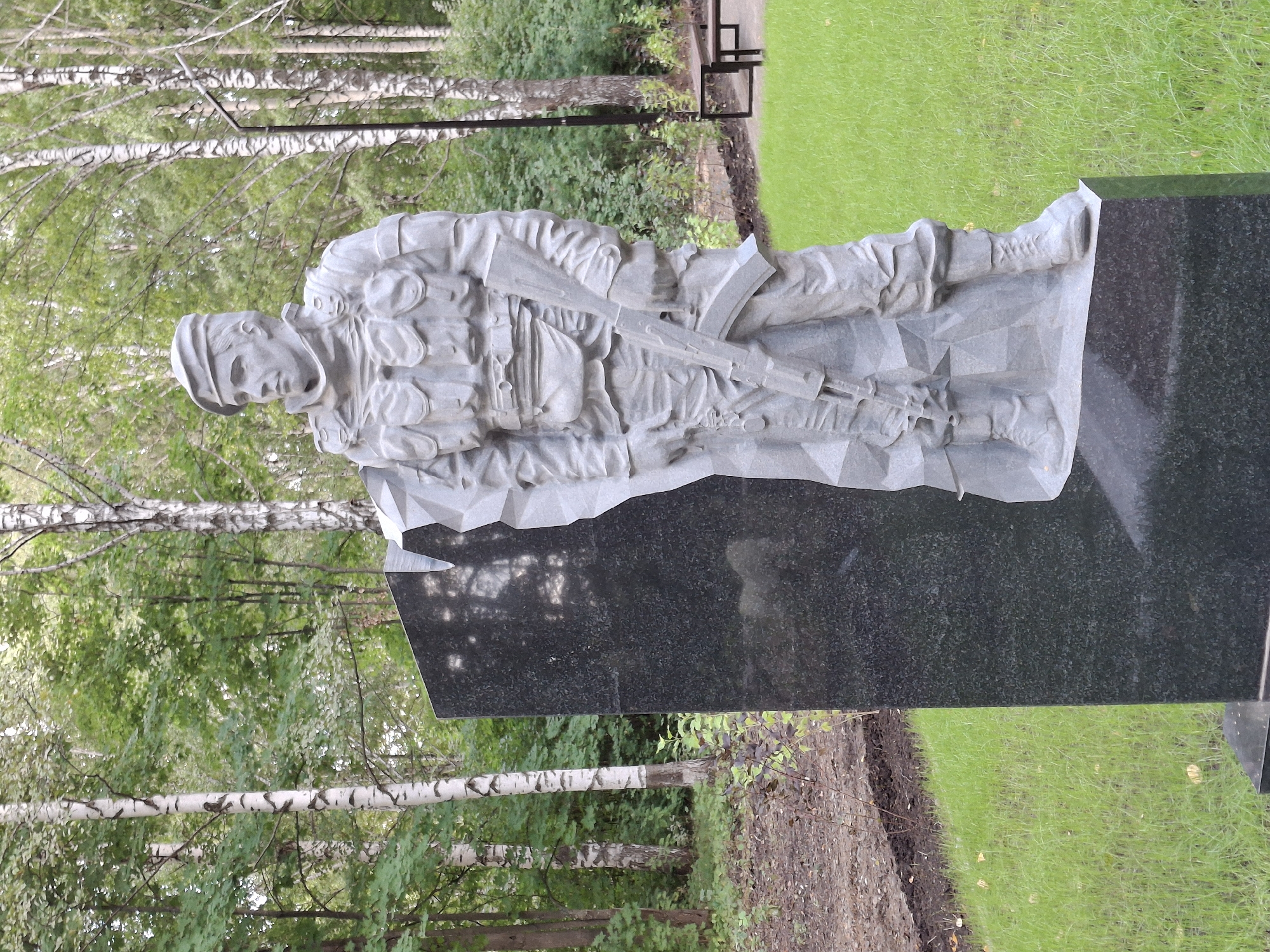 The image shows a stone sculpture of a soldier standing in front of a dark granite monument. The soldier is depicted wearing a beret, a combat vest, and carrying a rifle. The sculpture is rendered in a stylized, faceted manner. The monument has a geometric, broken surface on its left side, where it meets the soldier. The scene is set outdoors in a park-like environment with green grass, trees, and fallen leaves on the ground. There are birch trees in the background. A dark, angled bench with a black metal frame is visible to the right of the sculpture, near a paved area. The image is taken from a medium-low angle, focusing on the monument and the sculpture. The location is identified as Elovo, Russia, suggesting this is a war memorial or monument within a public space.