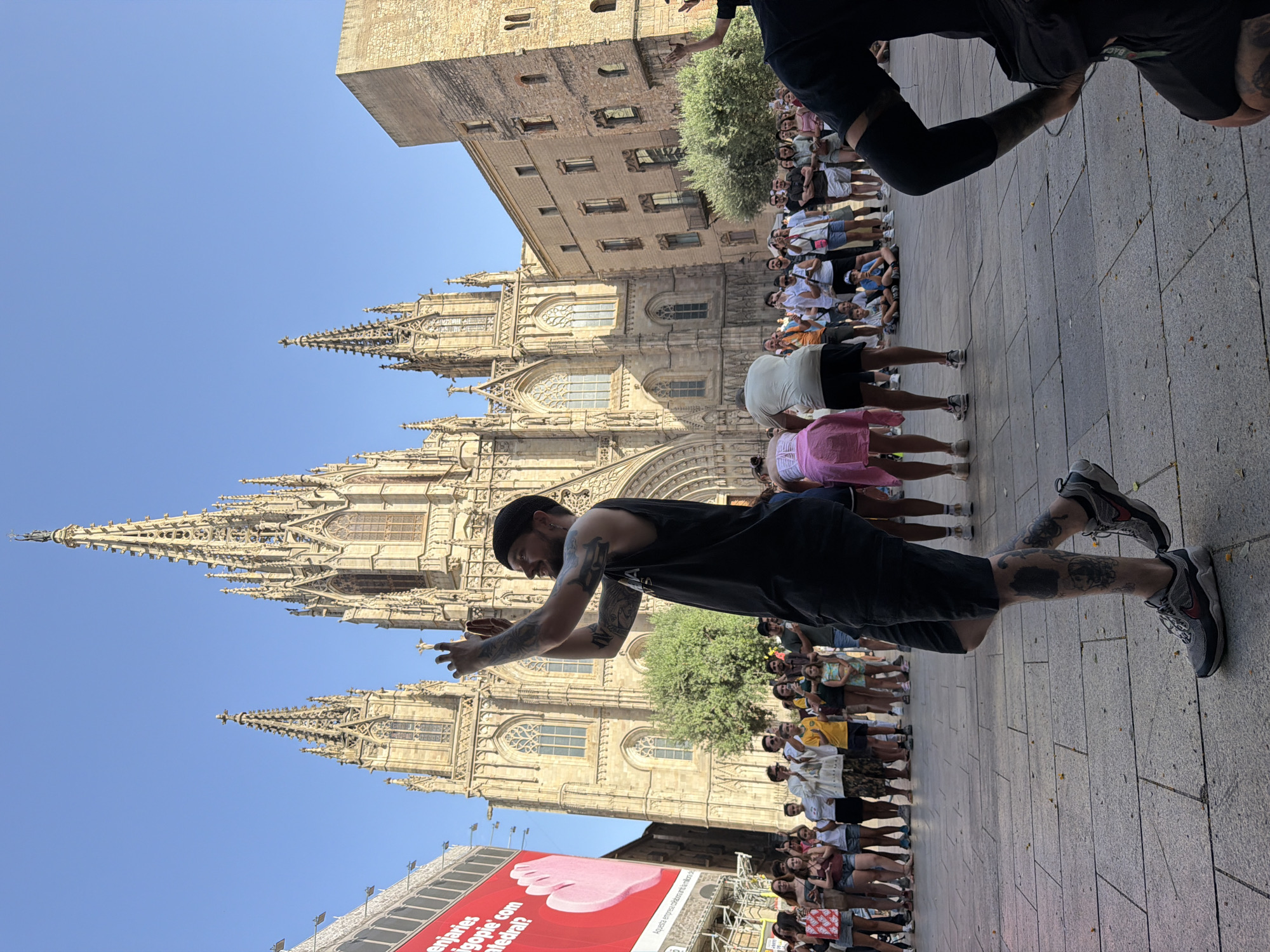 In Barcelona, Spain, in front of the Barcelona Cathedral, a crowd of people gathers on a paved plaza. A man in the foreground, facing away from the camera and toward the cathedral, is filming with his phone. He has tattoos on his arms and legs and is wearing a black tank top and shorts. Several other people are visible in the background, some sitting and some standing, watching the scene. Behind them, the ornate facade of the Barcelona Cathedral rises majestically against a clear blue sky. To the left of the cathedral, a large red banner with white text is displayed on a building. The overall scene suggests a lively public gathering, possibly a performance or event, in a historically significant location.
