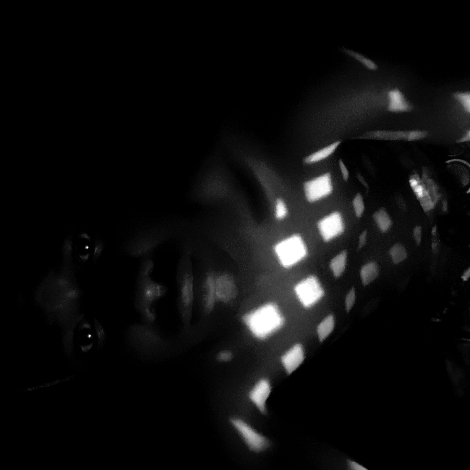 A black and white photograph depicts a person with dark skin lying down and looking towards the camera. The subject is partially obscured by a pattern of square light patches, possibly projected from an unseen source. The person's face is visible, featuring their eyes, nose, and mouth. The environment is dark, with only portions of the person's body illuminated. It's impossible to determine a location in Narayi, Nigeria from the available visual data.