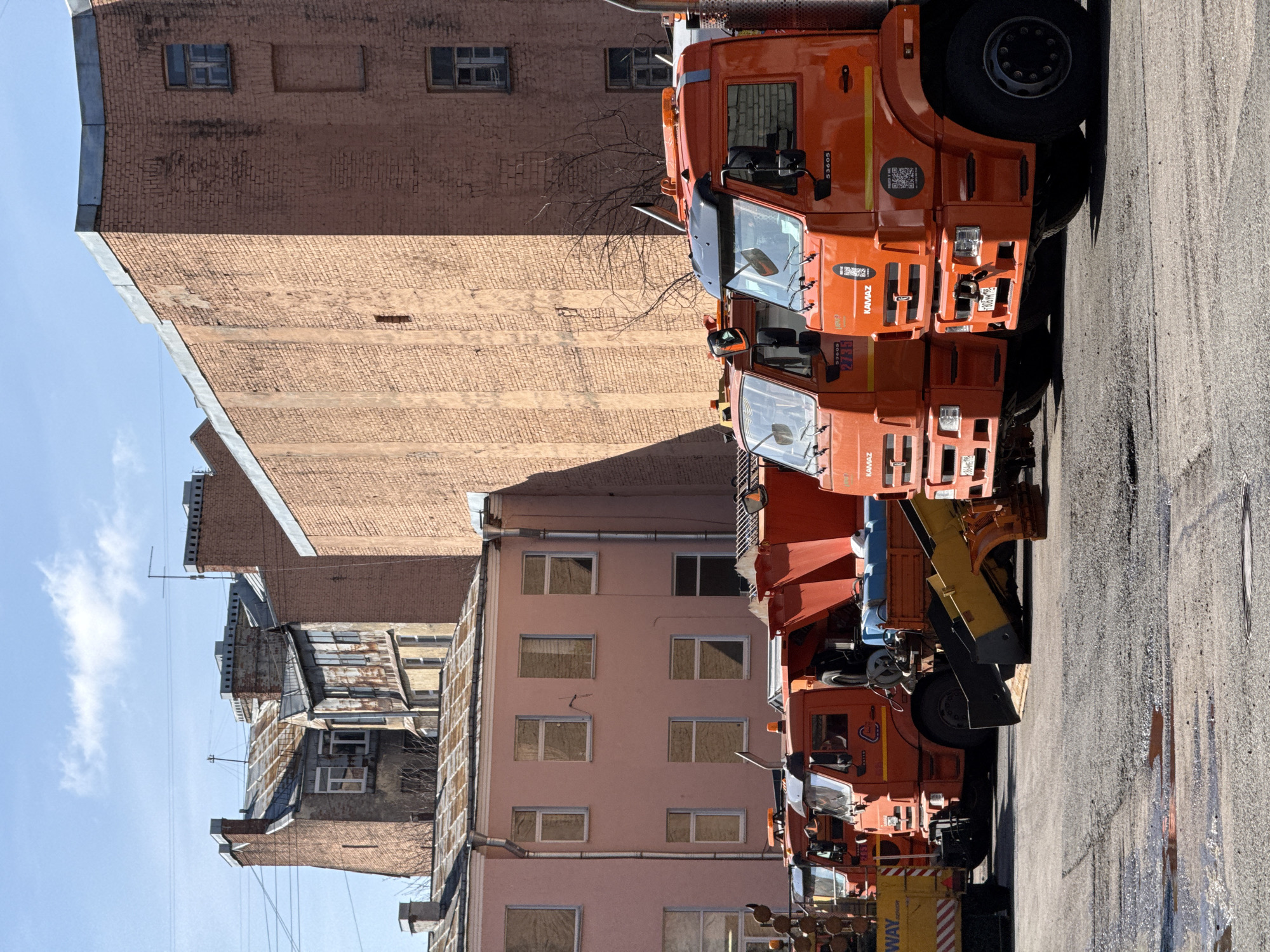 The image depicts a street scene in Saint Petersburg, Russia, showing several orange Kamaz trucks parked in a row along the roadside. The trucks appear to be utility vehicles, possibly for snow removal or road maintenance, given their configuration. The background consists of old brick buildings with various architectural styles, suggesting a historic urban area. The street itself looks wet, indicating recent rain or street cleaning. The sky is visible in the upper left corner, appearing clear with some clouds. The angle of the photograph indicates that the image was captured from the ground level, looking upwards at both the vehicles and the buildings. There are no people visible in the image.