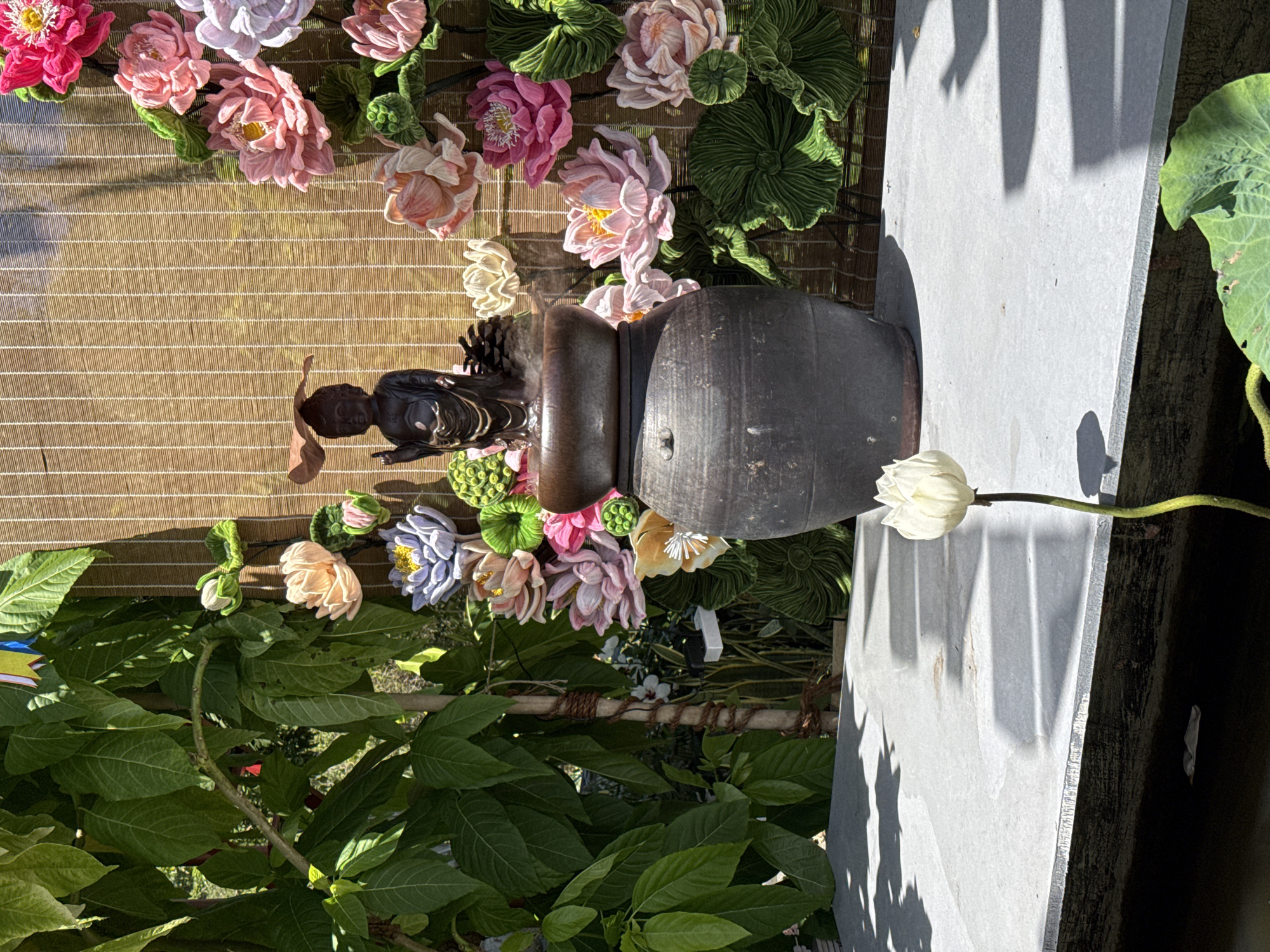 In Bac Nha Trang Ward, Vietnam, an outdoor scene features a stone statue positioned near a water jar and lotus flower. The statue is a dark hue. The water jar is cylindrical, rustic, and dark in color. A single, white lotus flower rests nearby. The background is decorated with artificial flowers in varying shades of pink, as well as green leaves. A woven backdrop hangs behind the statue and flowers. Green foliage surrounds the arrangement. A light-colored concrete slab with shadows is visible on one side.
