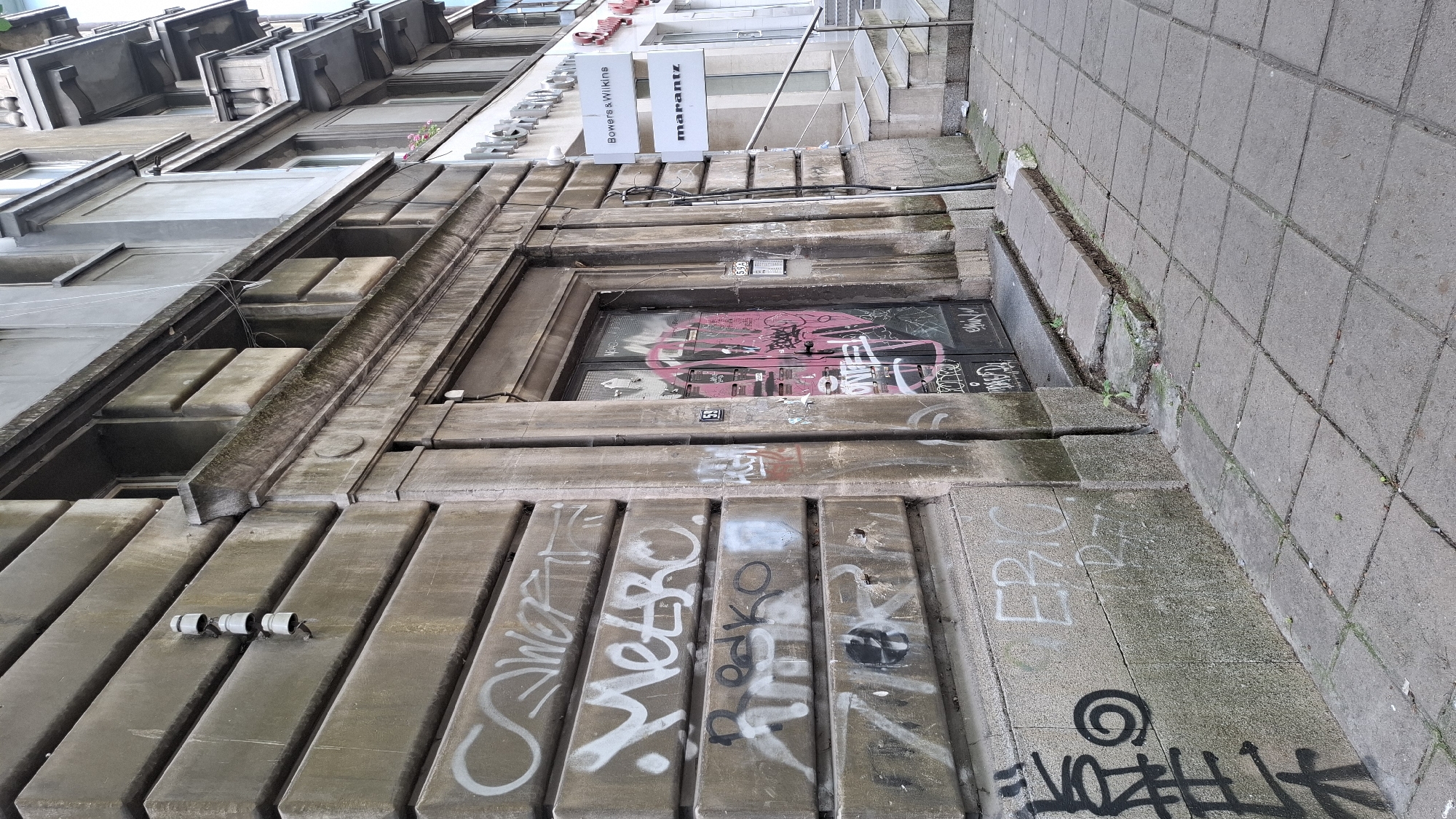 A low-angle, full shot captures the exterior of a weathered, Brutalist-style building in Sofia, Bulgaria. The building's facade is constructed from large, horizontal concrete blocks, heavily adorned with various forms of graffiti in white, black, and pink spray paint. The entrance to the building features a double door, also covered in graffiti, with a small sign displaying the number "59" above it. To the right of the entrance, three white signs are mounted on the building, two displaying brand names: "Bowers & Wilkins" and "marantz". A set of concrete stairs leads up to the building on the right side. The ground in front of the building is paved with square concrete tiles, some of which are cracked or displaced. The overall scene suggests an urban environment with signs of age and decay, contrasted with commercial signage.
