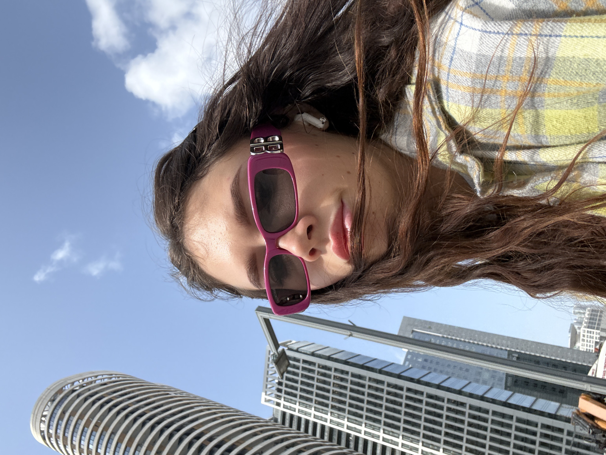 A low-angle shot captures a woman wearing pink sunglasses with dark lenses and an AirPod in her ear. She is wearing a white, green, and yellow plaid top. The background features the clear blue sky with a few small clouds and a cityscape dominated by modern buildings. A cylindrical building with horizontal lines is partially visible, along with a taller, rectangular building with a grid-like facade of windows. The location is Tel-Aviv, Israel.