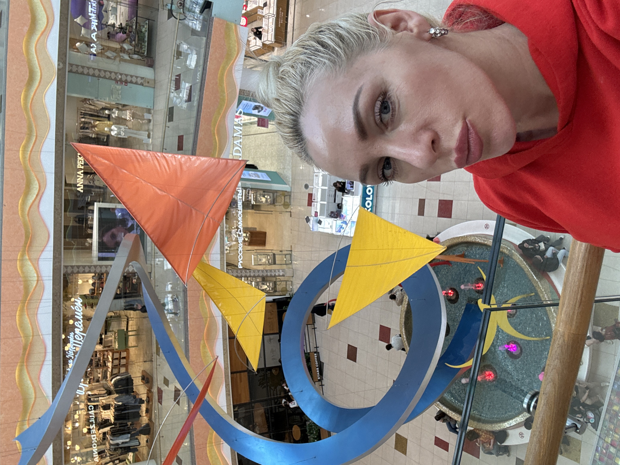 A woman with blonde hair in a high bun and wearing a red hoodie takes a selfie in a shopping mall in Unknown, Unknown. She is positioned at an elevated viewpoint overlooking the mall's central area. The background features a large decorative installation with colorful triangular shapes and a blue circular structure. There is a fountain below the structure, surrounded by people sitting and standing. Storefronts line the perimeter of the mall, and various signs in what appear to be Russian Cyrillic script are visible. The mall's interior is well-lit, with a mix of architectural details and modern design elements.