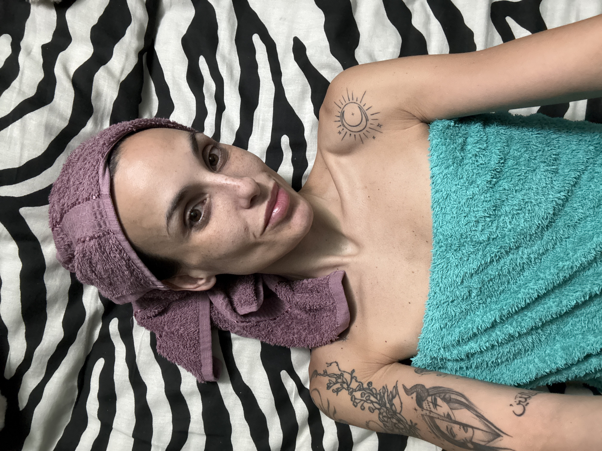 A person is lying on a zebra-striped bed. The person has a purple towel wrapped around their head and a teal towel draped across their body. The person has tattoos on their left shoulder and arm. The location is not discernible from the image.