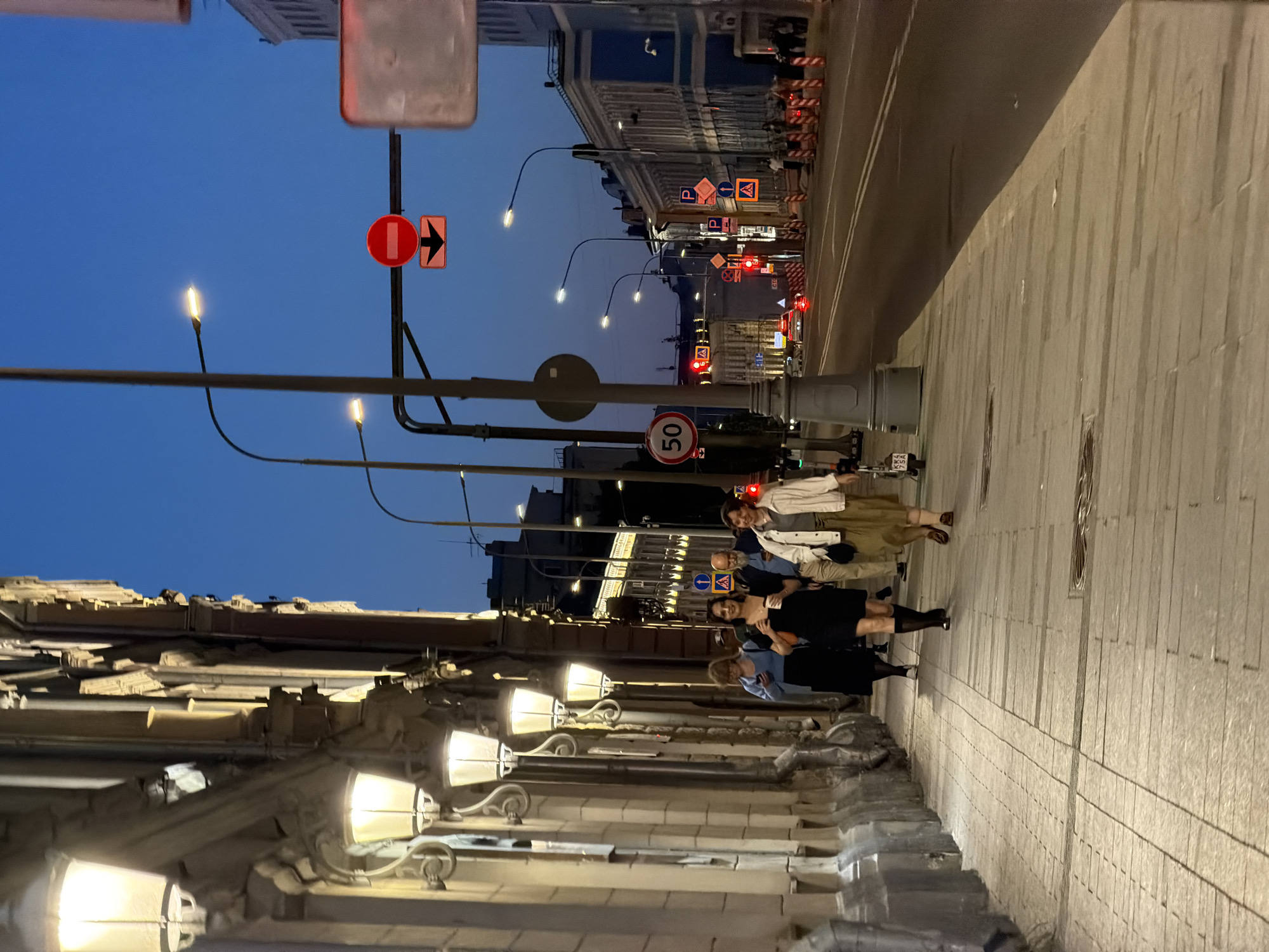 The image depicts a group of four people walking on the sidewalk of a city street in Moscow, Russia, at dusk. The sidewalk is paved with rectangular stone slabs. On the left side of the image is a building facade with ornate architectural details and illuminated street lamps. The street has a row of streetlights and various road signs, including speed limit and parking signs. The sky is transitioning from daylight to night.