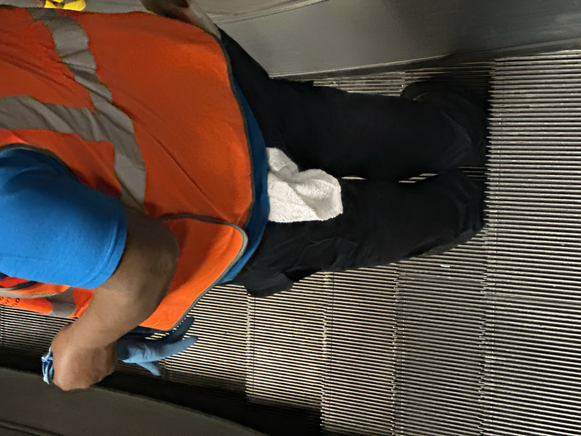 An individual wearing a blue t-shirt, an orange and grey safety vest, and black pants is on an escalator. The person has blue gloves on their hand that is visible. They also have a white towel on their pants. The escalator is made of metal slats. The location is in London, United Kingdom.