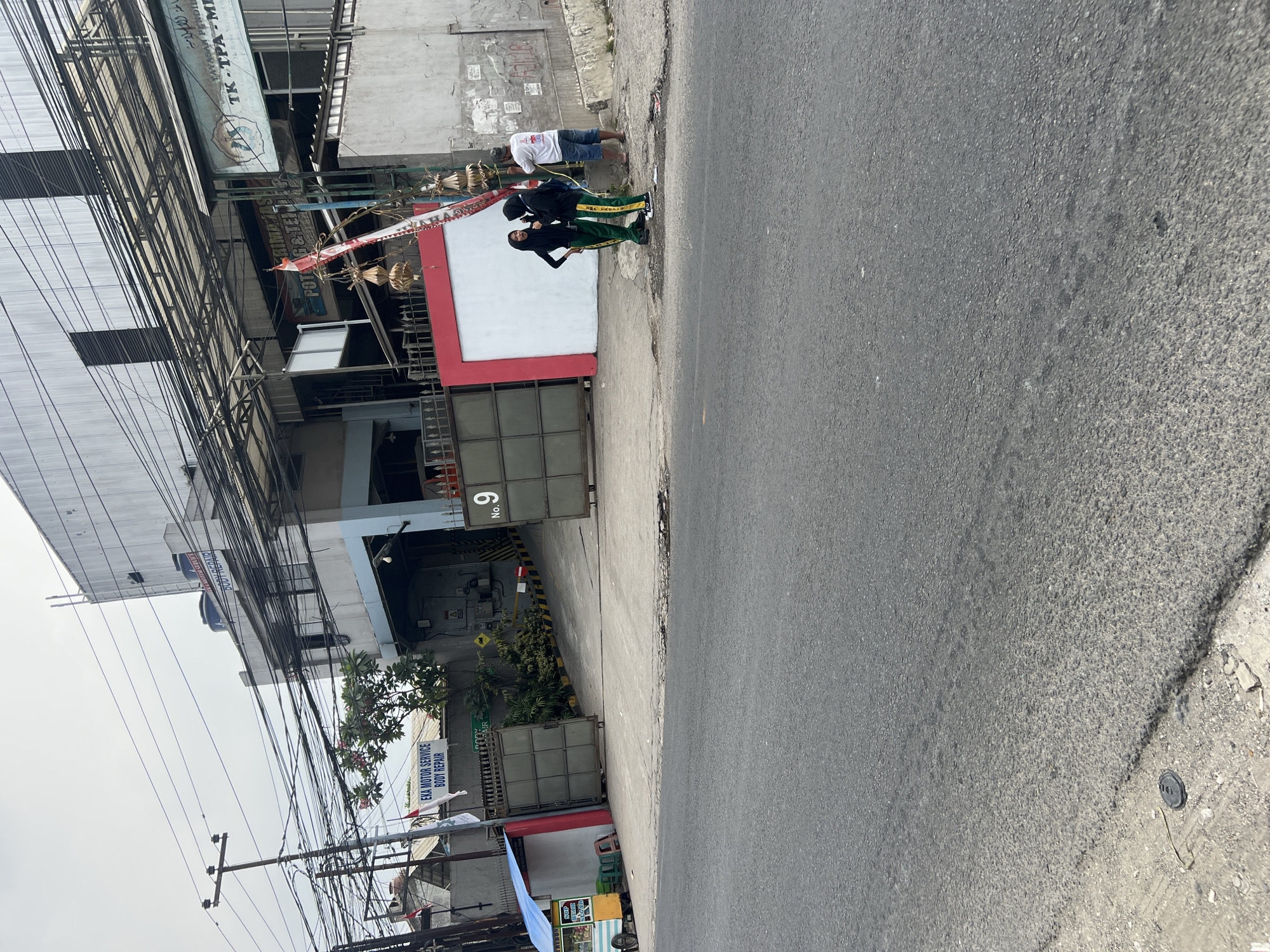 The image depicts a street scene in Citeureup, Indonesia. In the foreground, a gray asphalt road is visible, with a narrow concrete sidewalk to the right. Along the sidewalk, there are three individuals: two young women dressed in hijabs and green and yellow tracksuits, engaged in conversation, and an adult male wearing a white t-shirt and blue shorts, standing nearby. Behind the sidewalk, a building complex with a corrugated metal roof and light-colored walls is prominent. A large sign on the building reads "EKA MOTOR SERVICE BODY REPAIR". The complex features metal gates, some with a red frame, and a ramp leading into the property. Electrical wires are strung overhead, forming a dense network. To the left of the building entrance, a small cart selling fruits and drinks is parked, with two green plastic stools beside it. The environment appears to be commercial or industrial, with multiple signs and gates indicating various businesses. The sky is overcast.