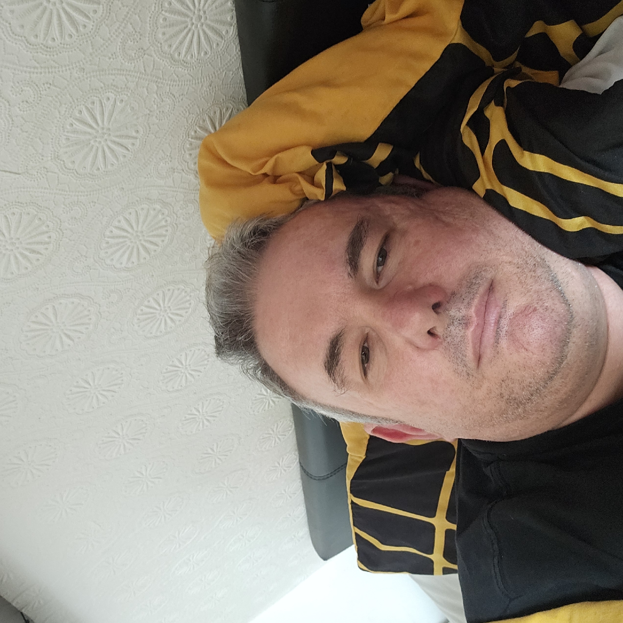 A middle-aged man with grey hair and light stubble is depicted lying down indoors in Holywell, United Kingdom. He is looking directly towards the camera with a neutral or slightly contemplative expression. He is partially covered by or wearing a black garment featuring prominent yellow stripes and patterns, suggesting it could be a jacket, blanket, or similar attire. Behind his head, a dark, possibly leather, surface is visible, likely a headrest or cushion. The dominant background is a white, textured surface, possibly wallpaper or a bedspread, adorned with a repeating embossed pattern of stylized flowers or stars enclosed within circles. The lighting is bright and diffused, indicating an indoor setting with ample illumination. No text is visible in the image.