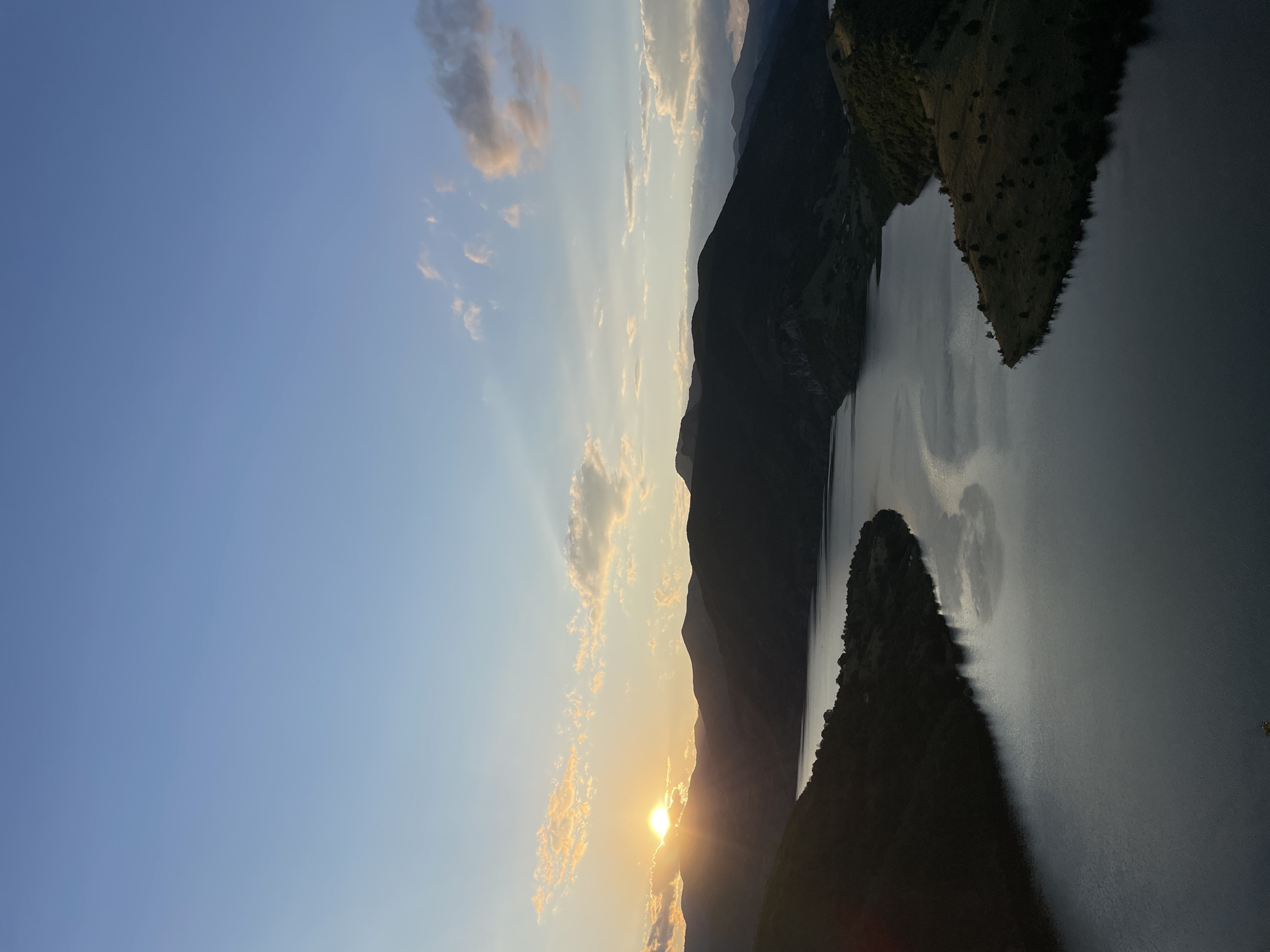 This is an aerial, high-angle view of a body of water, likely a lake or reservoir, situated amidst a mountainous landscape. The sun is setting behind a range of dark, silhouetted mountains on the left side of the image, casting a warm glow on the sky and some scattered clouds. The water itself is calm and reflects the twilight sky and the surrounding hills. The terrain on the right side of the frame shows rolling hills with sparse vegetation and trees, suggesting a rural or natural environment. The overall scene conveys a sense of peacefulness and natural beauty during dusk. The location is identified as Borovitsa, Bulgaria.