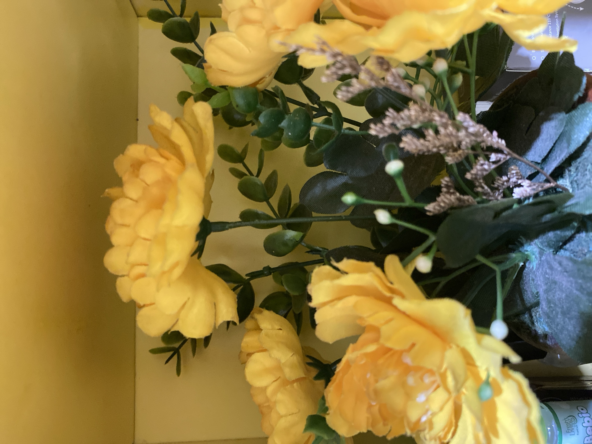 A close-up, eye-level shot captures a bouquet of artificial yellow flowers with green leaves and smaller, dried filler flowers. The background consists of a plain yellow wall, suggesting an indoor setting. The focus is on the vibrant colors and textures of the floral arrangement. The image does not provide any information about the location in Lambaro Kueh, Indonesia, or any people or actions.