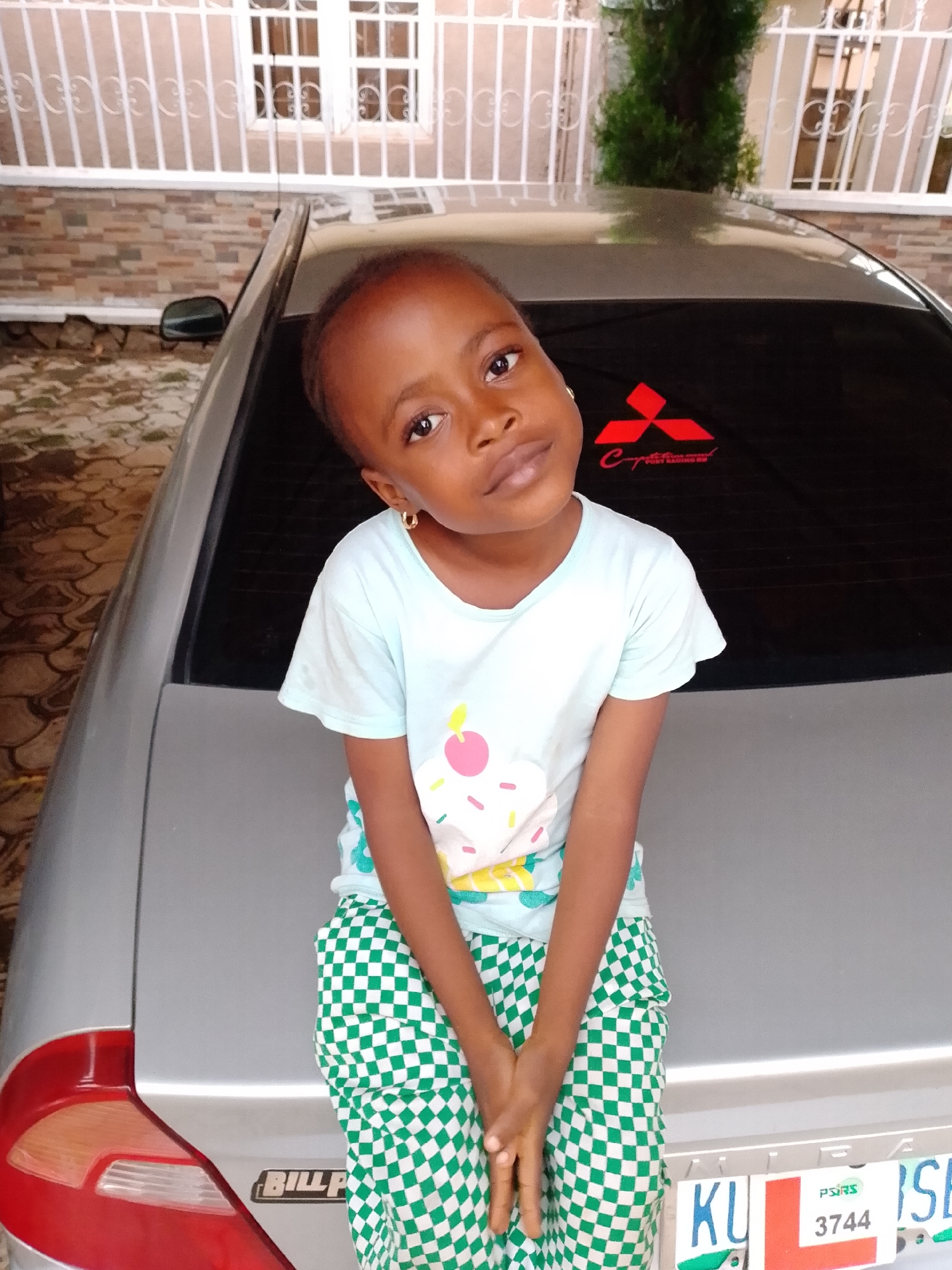 A young girl with dark skin, wearing a light blue t-shirt with a cupcake design and green and white checkered pants, sits on the trunk of a silver car. She is looking at the camera with her head tilted to the left and her hands clasped in front of her. A Mitsubishi logo sticker is visible on the car's rear window. The background shows a brick wall and a white-railed structure. The scene appears to be outdoors, possibly in a residential area.