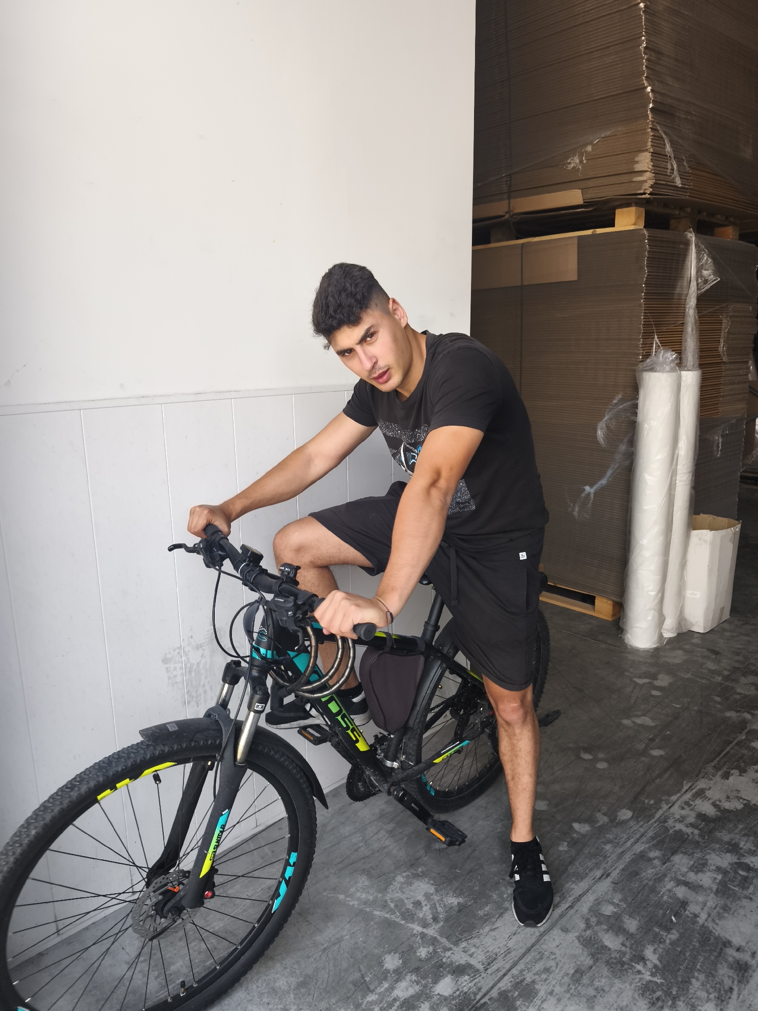 A young man with short, dark, curly hair, wearing a black t-shirt and black shorts, is sitting on a black and green mountain bike. He is leaning forward, with his left hand on the handlebars and his right arm bent at the elbow, resting on his thigh. His gaze is directed towards the camera. The bicycle has black tires with yellow and blue accents, and a black saddlebag. The man is positioned in front of a white wall with horizontal paneling on the left side of the frame. To the right of the man and the bicycle, there are tall stacks of cardboard boxes wrapped in plastic, and several large white rolls of material. The floor is concrete, showing signs of wear and tear. The background suggests an indoor industrial or storage environment.