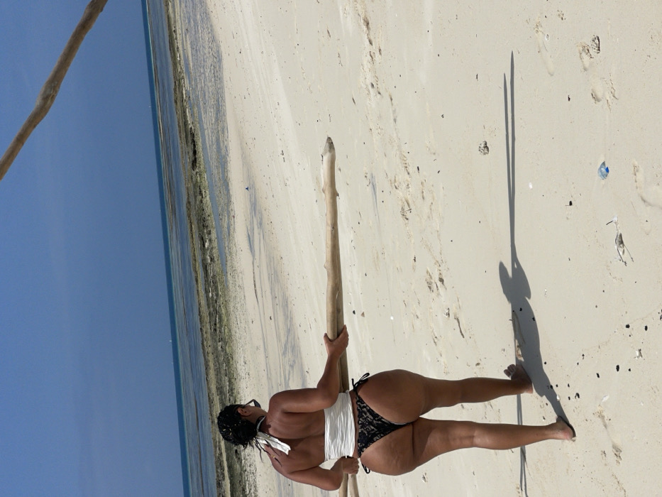 The image displays an outdoor scene on a sandy beach in Zanzibar, Tanzania, under a clear blue sky. An adult female is depicted from the back, walking away from the viewer towards the ocean. She wears a white halter-neck top and black patterned thong-style bikini bottoms with side ties. Her dark hair is pulled back, and sunglasses are positioned on her neck. She is barefoot and carries a long, light-colored piece of driftwood horizontally across her lower back, held by both hands. Her shadow, along with the shadow of the driftwood, extends prominently on the sand to the right. The beach consists of light-colored sand with scattered small debris, including what appears to be a blue plastic bottle cap. Beyond the immediate sandy shore, a wide expanse of rocky, exposed reef is visible, leading to the deeper blue water of the ocean. A faint, distant object, possibly a boat, is discernible on the horizon. A second, darker piece of wood or branch is partially visible in the upper right corner of the frame, extending diagonally.