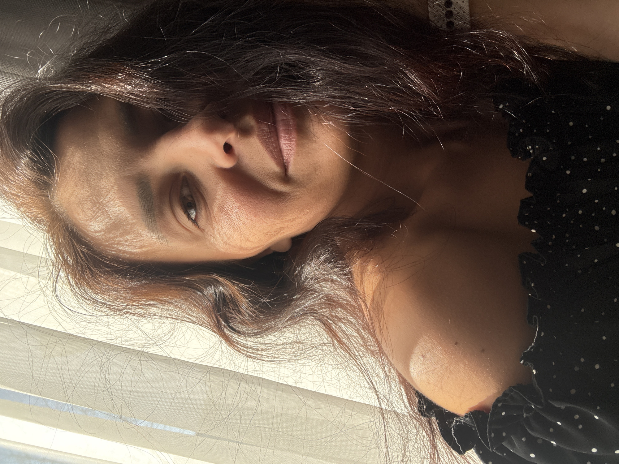 The image depicts a woman with brown hair lying down, possibly indoors. She is wearing a dark polka-dot top. Sunlight filters through a white, semi-transparent curtain. The location of the image is not determined. The location of Chaikovsky, Russia is not determined from this image.