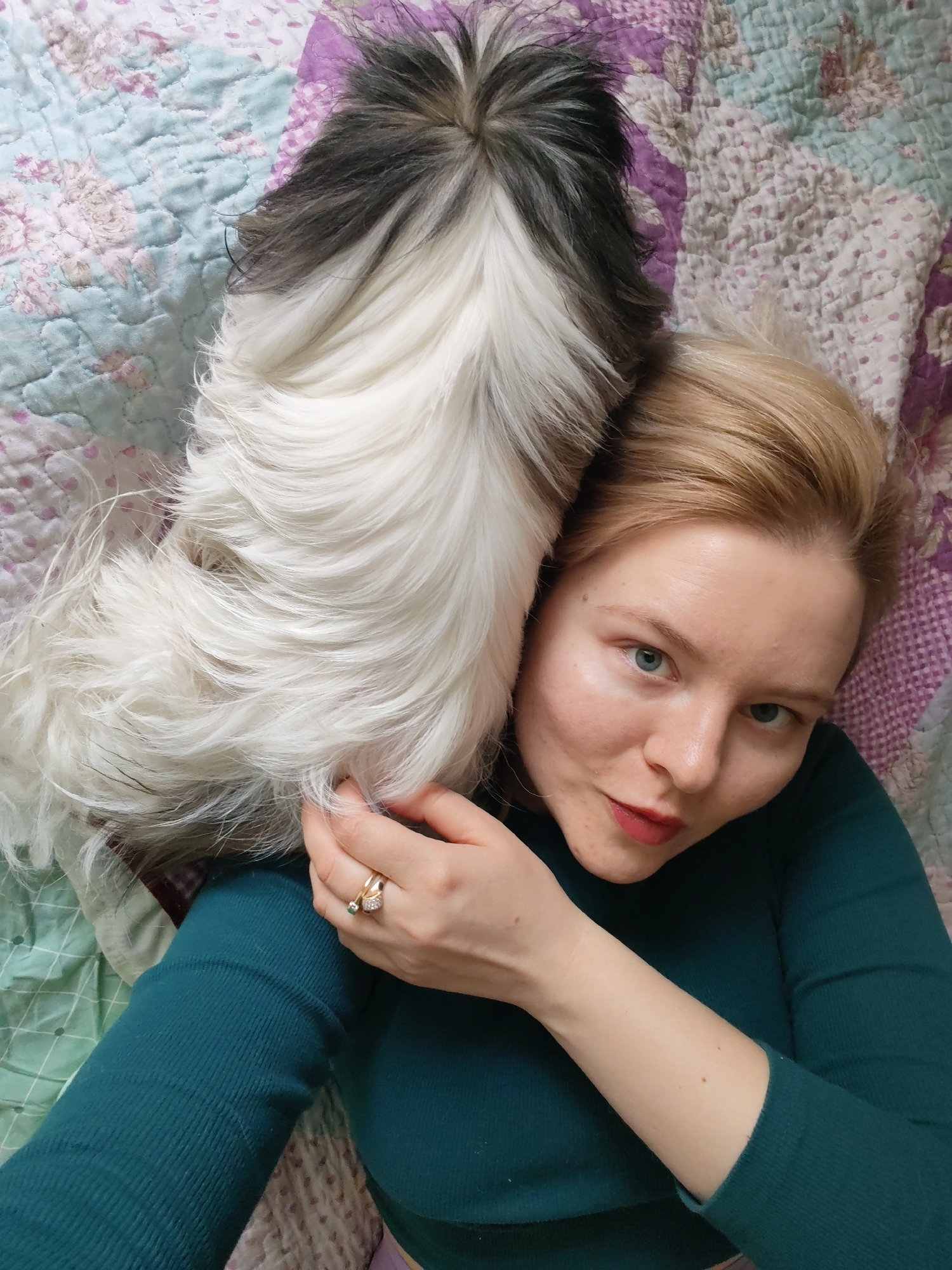 An eye-level, close-up shot shows a fair-skinned woman with blonde hair and blue eyes lying down next to a fluffy dog, likely indoors on a bed or couch. The woman is positioned on the right side of the frame, looking directly at the camera with a calm expression and a hint of red lipstick. She is wearing a teal long-sleeved top, and a golden ring with a green stone is visible on the ring finger of her right hand, which is resting near the dog's fur.
The dog, a small, long-haired breed, occupies the left side of the frame, snuggled closely to the woman's head and shoulder. Its fur is predominantly white and very thick, with patches of black and gray visible on its back and head. The dog's head is turned away from the camera, resting beside the woman's head.
They are both lying on a quilted bedspread or blanket that features a patterned design with various shades of pink, purple, light blue, and white. The lighting is soft and bright, suggesting daytime. The overall scene conveys a cozy, affectionate moment between a pet owner and her dog, captured in Izhevsk, Russia. There is no visible text in the image.