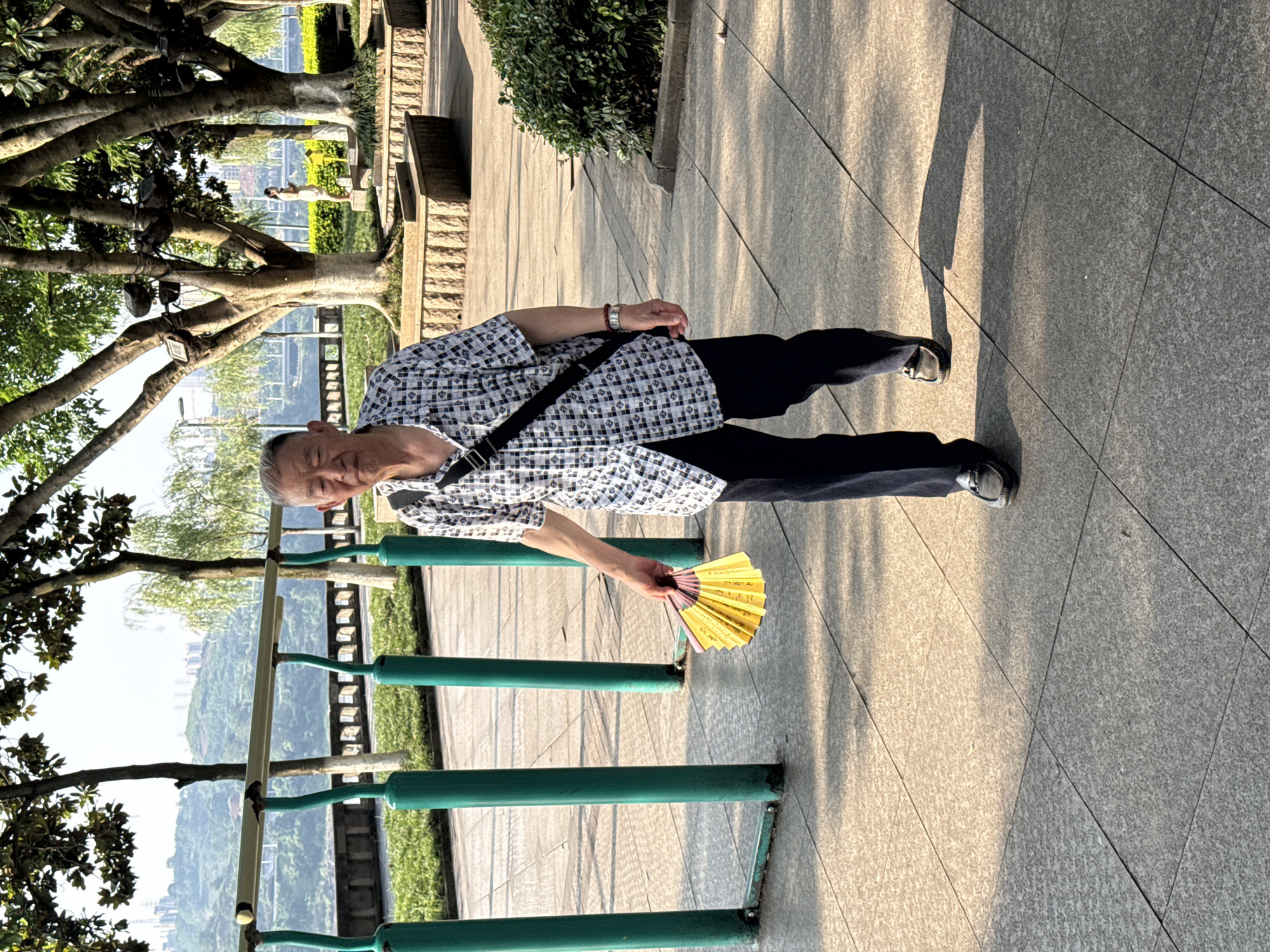 An older East Asian man is the central subject of the image, standing on a paved outdoor walkway in what appears to be a park or scenic area. He has short, grey hair and is wearing a short-sleeved, patterned shirt in black and white, dark trousers, and dark slip-on shoes. A black cross-body bag is slung over his right shoulder, and a watch is visible on his left wrist. He holds a yellow folded fan in his right hand, which has some faint, dark writing on it that is not clearly legible.
The man is looking towards the viewer with a slight, pleasant smile, suggesting a relaxed mood. He is standing under clear skies, indicated by the bright light and long shadows cast on the light gray paving stones, characteristic of either morning or late afternoon sun.
To the man's left, there are sturdy green metal railings, beyond which a verdant landscape unfolds. Large trees with thick trunks and green foliage frame the left side of the image, offering partial shade. In the background, beyond the railings and trees, a panoramic view of rolling green hills, dotted with distant buildings or structures, can be seen, hinting at an elevated location overlooking a valley or city. A small, manicured bush is visible on the right side of the path, further contributing to the park-like setting.