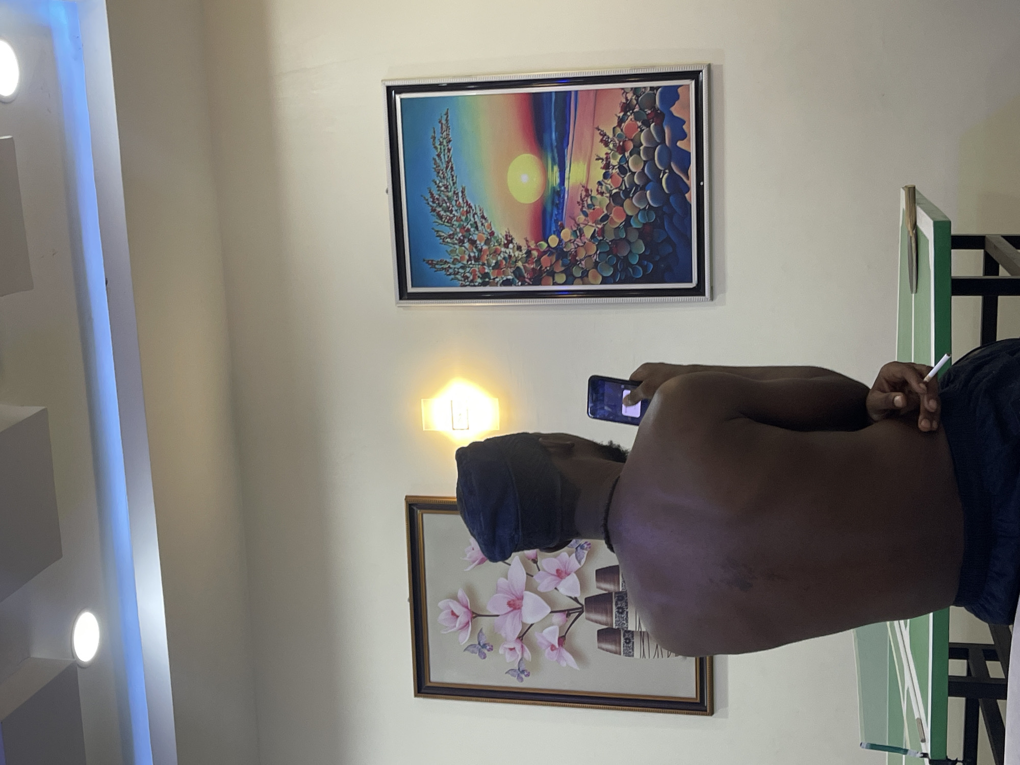 An indoor photograph, taken in Gwarinpa, Nigeria, captures a room with light-colored walls adorned with artworks and various lighting. A person, viewed from behind and facing away from the camera, stands towards the right of the frame. They have dark skin, are bare-chested, and wear a dark cap and dark shorts. The person is holding a smartphone in their right hand, appearing to capture an image or video, and also holds a white cylindrical object, possibly a cigarette, between their fingers.
On the wall above the person, a large framed painting depicts a vibrant, abstract landscape featuring a bright central sun or moon surrounded by colorful, organic shapes resembling foliage or fruit against a rainbow-hued sky. Below a glowing, wall-mounted rectangular light fixture emitting a warm yellow light, a second framed artwork displays delicate pink magnolia-like flowers with two butterflies, set against a subtle background featuring what appears to be potted plants.
The room is further illuminated by modern artificial light sources, including cool blue LED strip lighting running along an architectural detail on the ceiling to the left, and bright white recessed circular lights embedded in the ceiling. To the right of the person, a partial view of a green and white structure, possibly furniture or part of a partition, is visible.