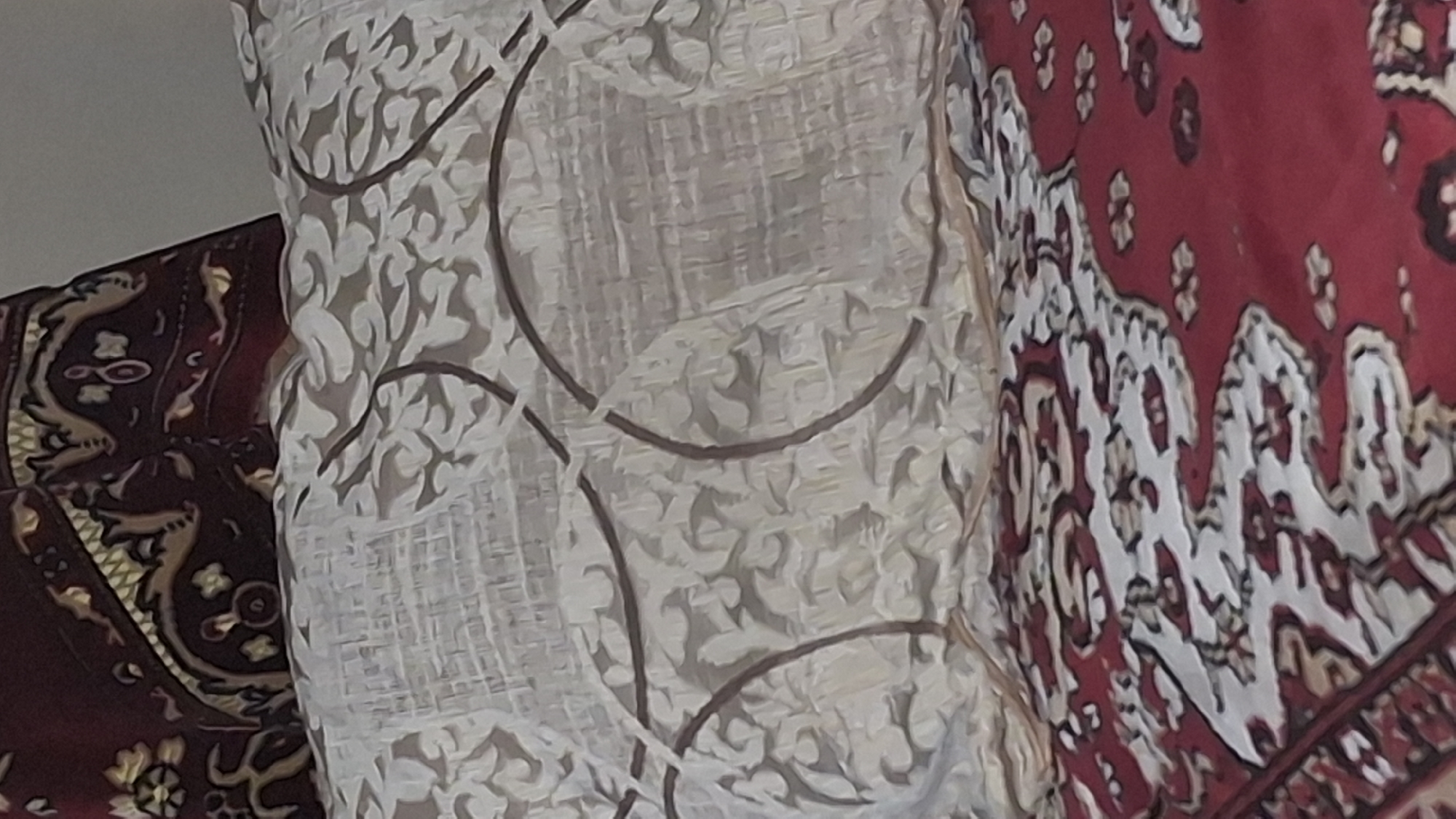 The image presents a close-up view of three distinct patterned textiles, possibly within an interior setting in Gyumri, Armenia. On the left, a section of burgundy fabric, likely a rug or upholstered furniture, features intricate traditional patterns in gold, beige, and dark brown, suggesting stylized floral or animal motifs. In the center, a light-colored textile, appearing off-white or beige, displays a layered design: a subtle, lighter patterned base is overlaid with prominent, darker brown abstract curved lines, some forming partial circles. This fabric could be a curtain or another piece of upholstery. On the right, another burgundy fabric is visible, distinct from the one on the left, showcasing a traditional carpet or rug design with bold white and lighter-toned patterns contrasting against its rich red background.
No people are visible, and no specific activity or event is taking place; the image is a still life focusing on the interplay of these decorative fabrics. The lighting is diffused, offering no information about the time of day. The traditional and elaborate patterns on the burgundy textiles are consistent with cultural designs found in the region, providing a strong contextual cue for the specified location of Gyumri, Armenia. There is no visible text in the image.