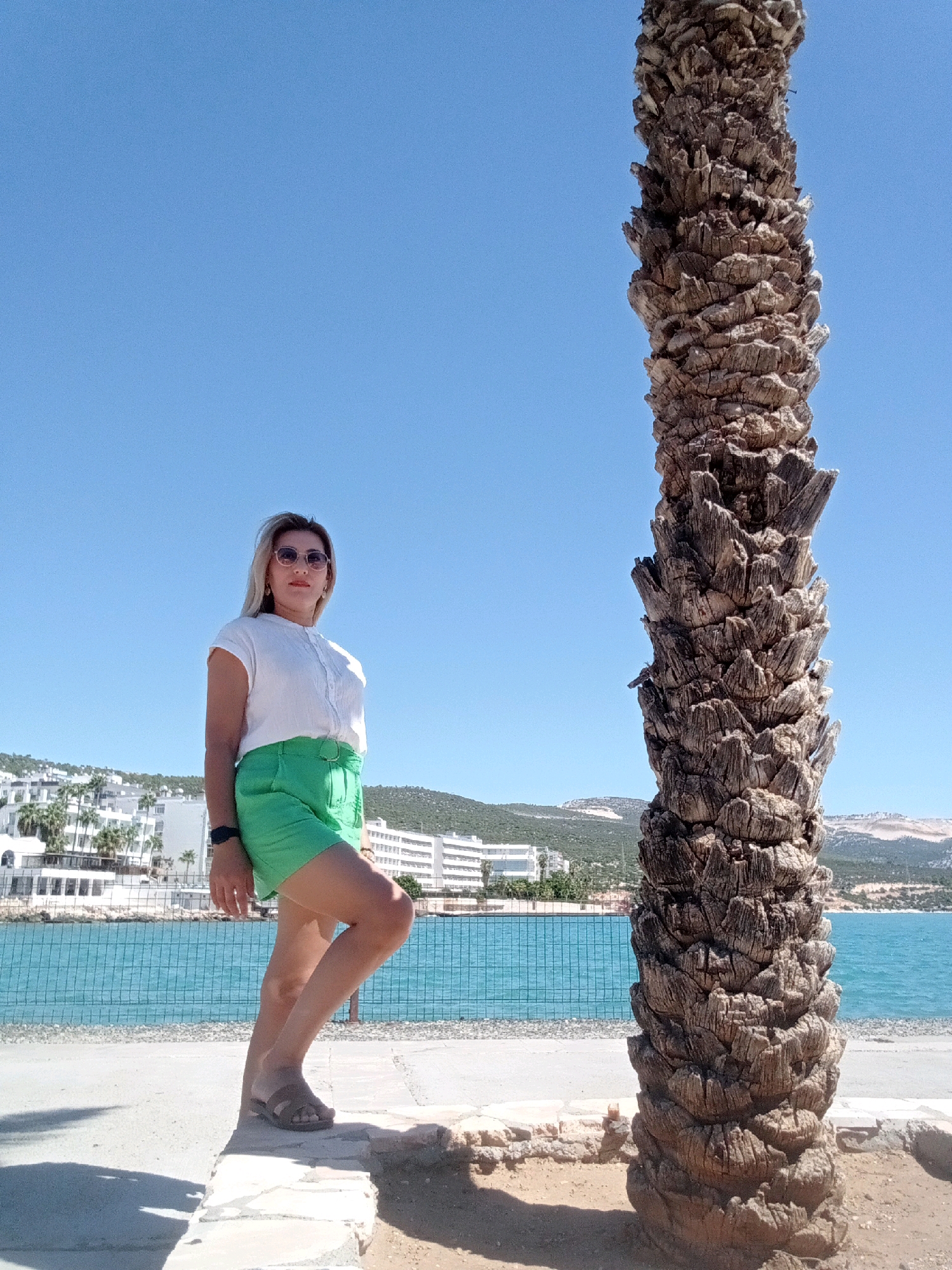 A woman stands in front of a large palm tree trunk with a clear blue sky. She is wearing a white short-sleeved shirt and green shorts, with sunglasses and a watch. The background shows a body of water, a fence, and buildings on a hill, suggesting a coastal city. The location is Silifke, Turkey.
