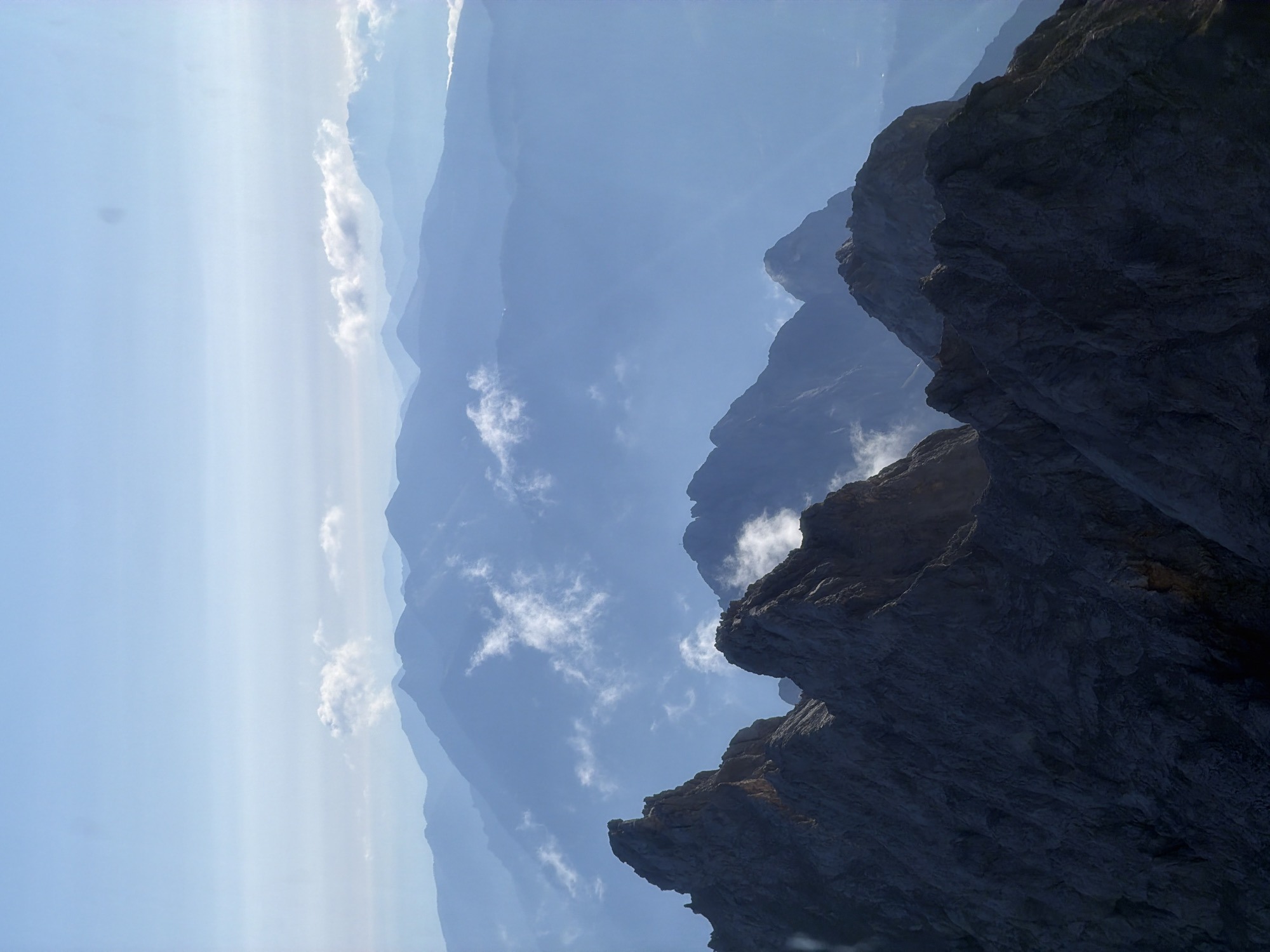 The image depicts a vast mountain range, seen from a high vantage point. The foreground is dominated by rugged, dark grey rock formations, characteristic of alpine peaks. Layers of mountains recede into the background, each appearing bluer and more indistinct due to atmospheric perspective. Wisps of white clouds are scattered throughout the mid-ground, clinging to the slopes and summits. The sky above is a pale, clear blue, with a faint hazy band near the horizon. The overall impression is one of immense scale and natural beauty. This scene is likely captured in the vicinity of Grainau, Germany, a municipality known for its proximity to the Wetterstein Mountains, which include Germany's highest peak, the Zugspitze, and feature dramatic rock formations and expansive alpine vistas. No people or artificial structures are clearly visible in the image, emphasizing the natural and untamed environment.