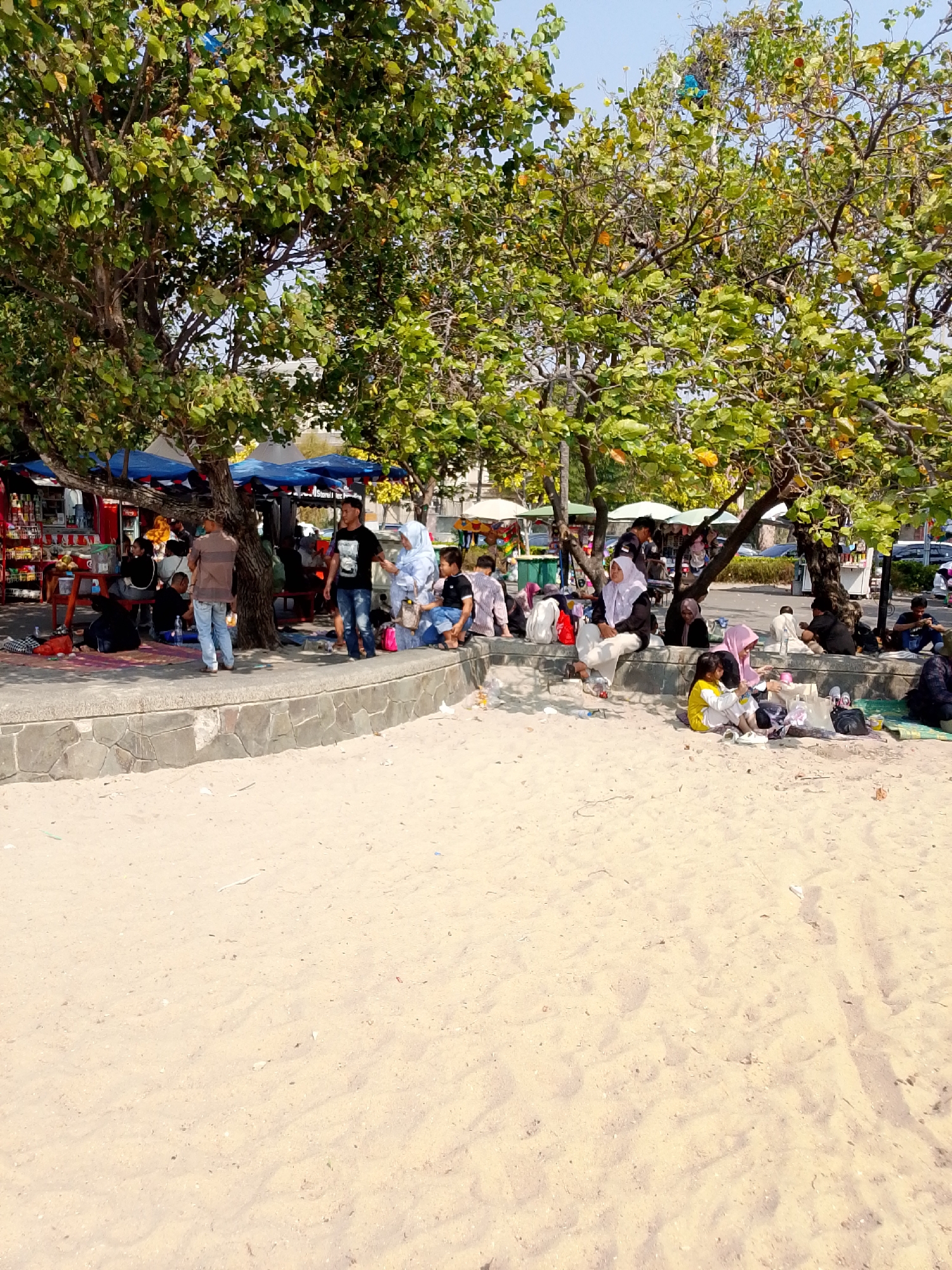 The image shows a crowded beach scene under large trees in Ancol, North Jakarta, Indonesia. Several people are seated on the sand or on a stone wall, many of them appearing to be families or groups of friends. Some are wearing casual clothing, while others, particularly women, are wearing hijabs. There are several stalls or kiosks visible behind the people seated on the wall, with colorful awnings and merchandise displayed. The foreground of the image is dominated by sand, with some scattered debris. The overall atmosphere suggests a lively public gathering place, possibly a tourist area or a popular local spot.