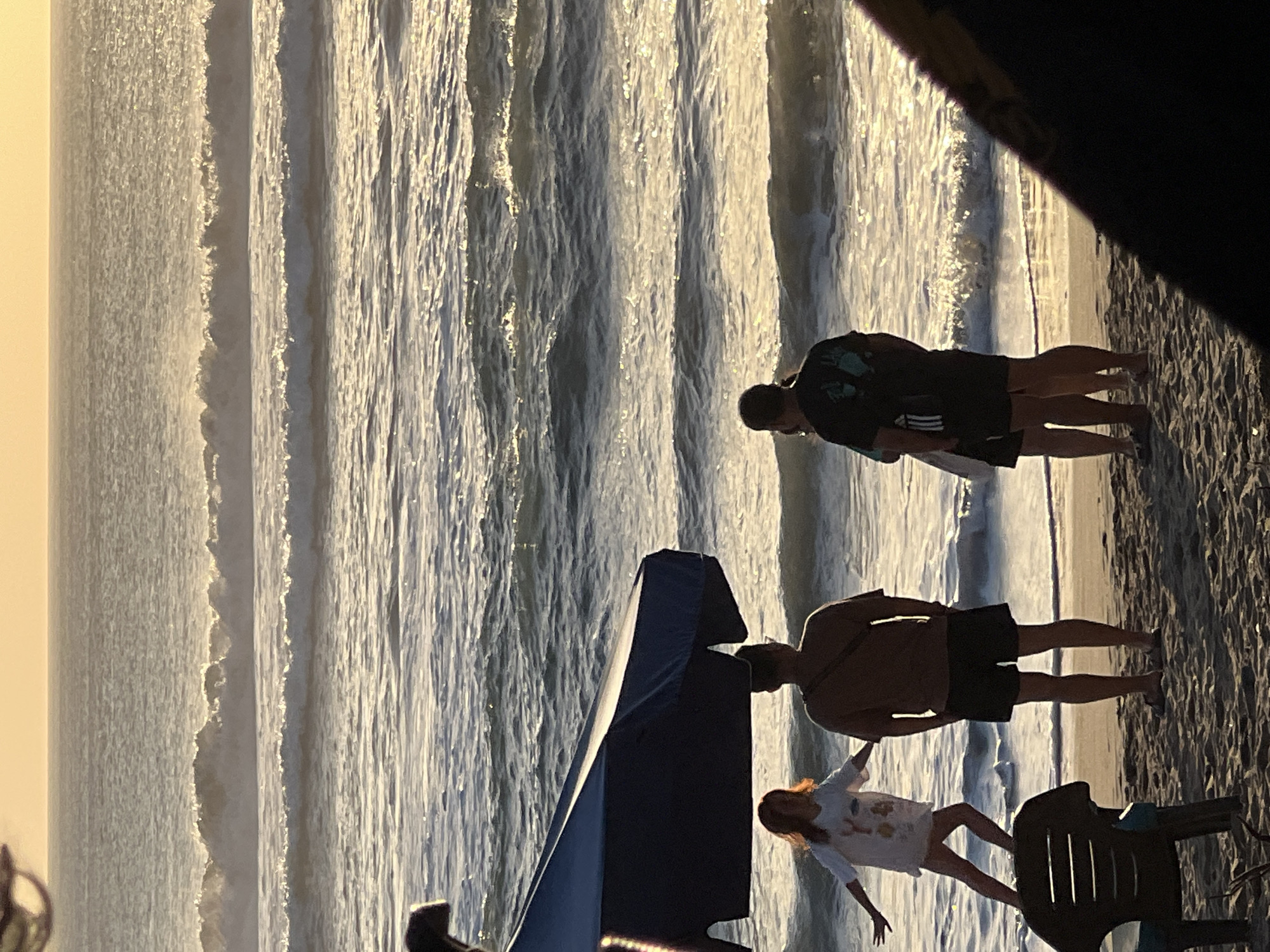 The image shows a beach scene at sunset in Kuta, Indonesia. The sun is setting, casting a warm glow over the ocean. Gentle waves are breaking on the shore. In the foreground, two adults stand near an umbrella. To the left, a young girl is playing on the sand. A beach chair is visible in the bottom left corner of the image.
