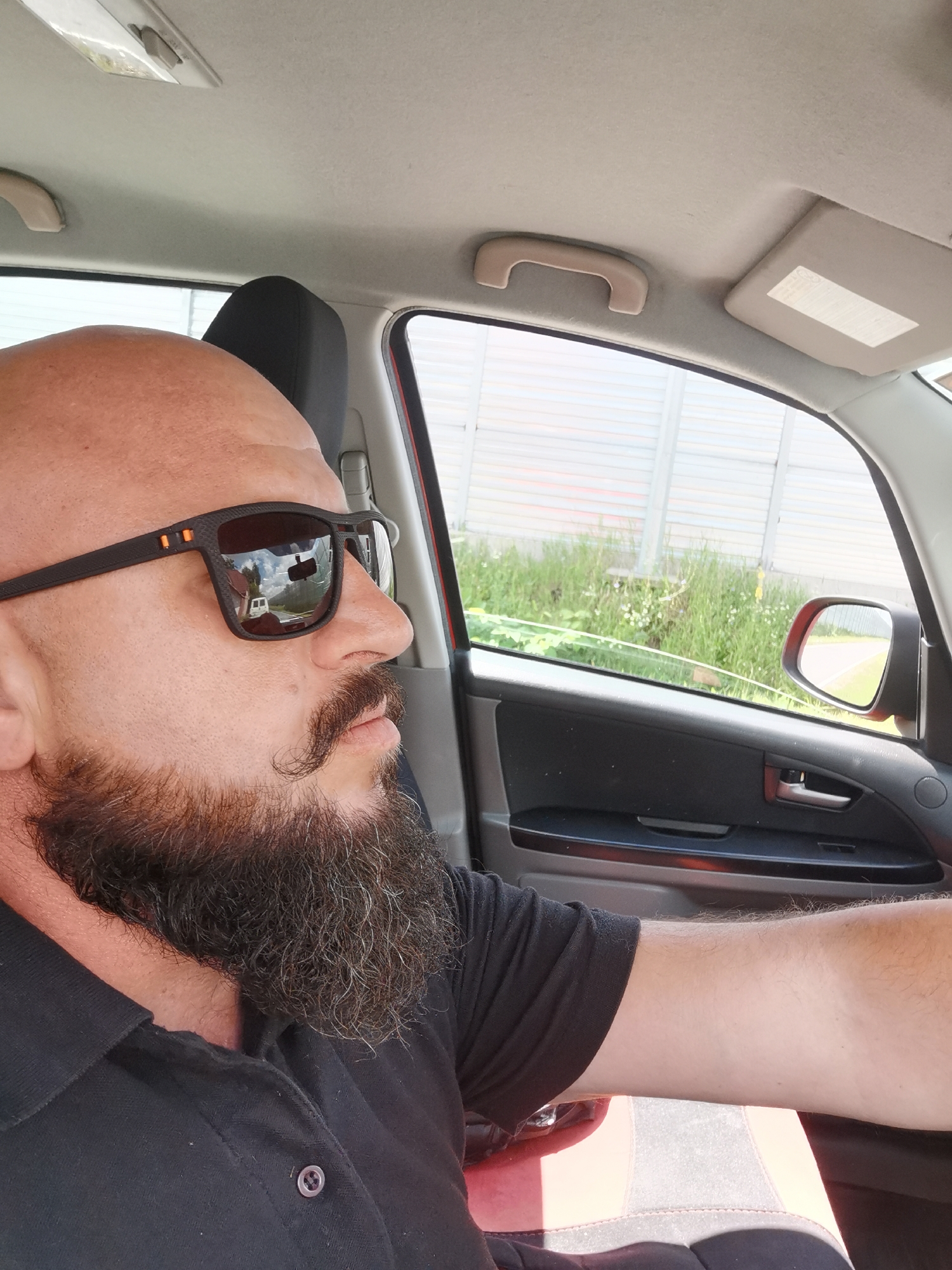 A bald man with a beard and mustache, wearing sunglasses and a black polo shirt, is driving a car. The interior of the car is visible, including the dashboard, door panel, and a headrest. Through the car window, a grassy roadside and a white fence are visible. The image appears to have been taken on a sunny day. The location is identified as Semypolky, Ukraine.