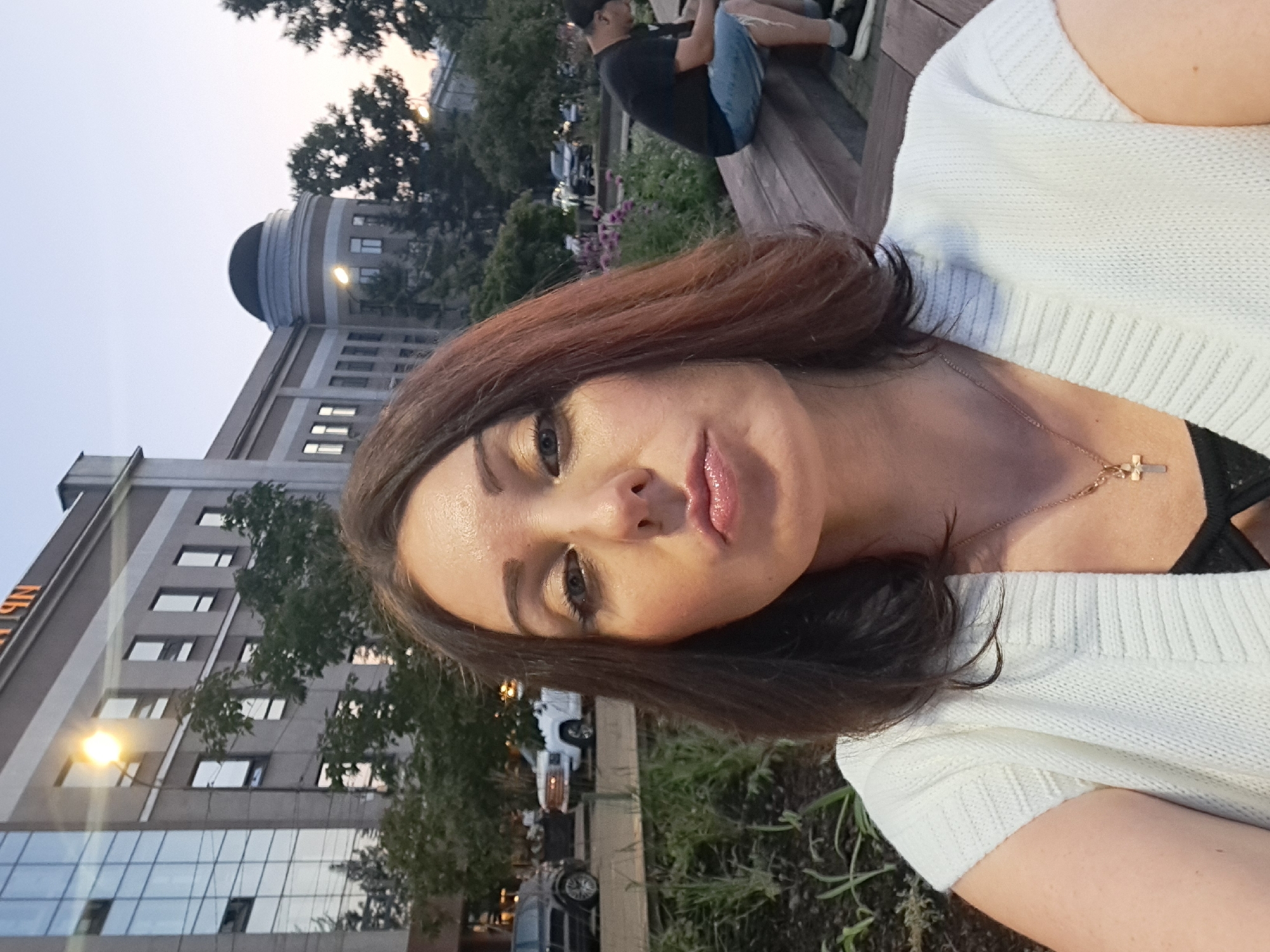 A woman with shoulder-length brown hair and makeup is taking a selfie outdoors. She is wearing a white knit sweater and a gold cross necklace. Behind her, a man in a black t-shirt and denim shorts is sitting on a wooden bench, looking at his phone. The background shows a modern building with glass windows, trees, and a street with cars. The sky is dusk. This scene is taking place in Irkutsk, Russia.